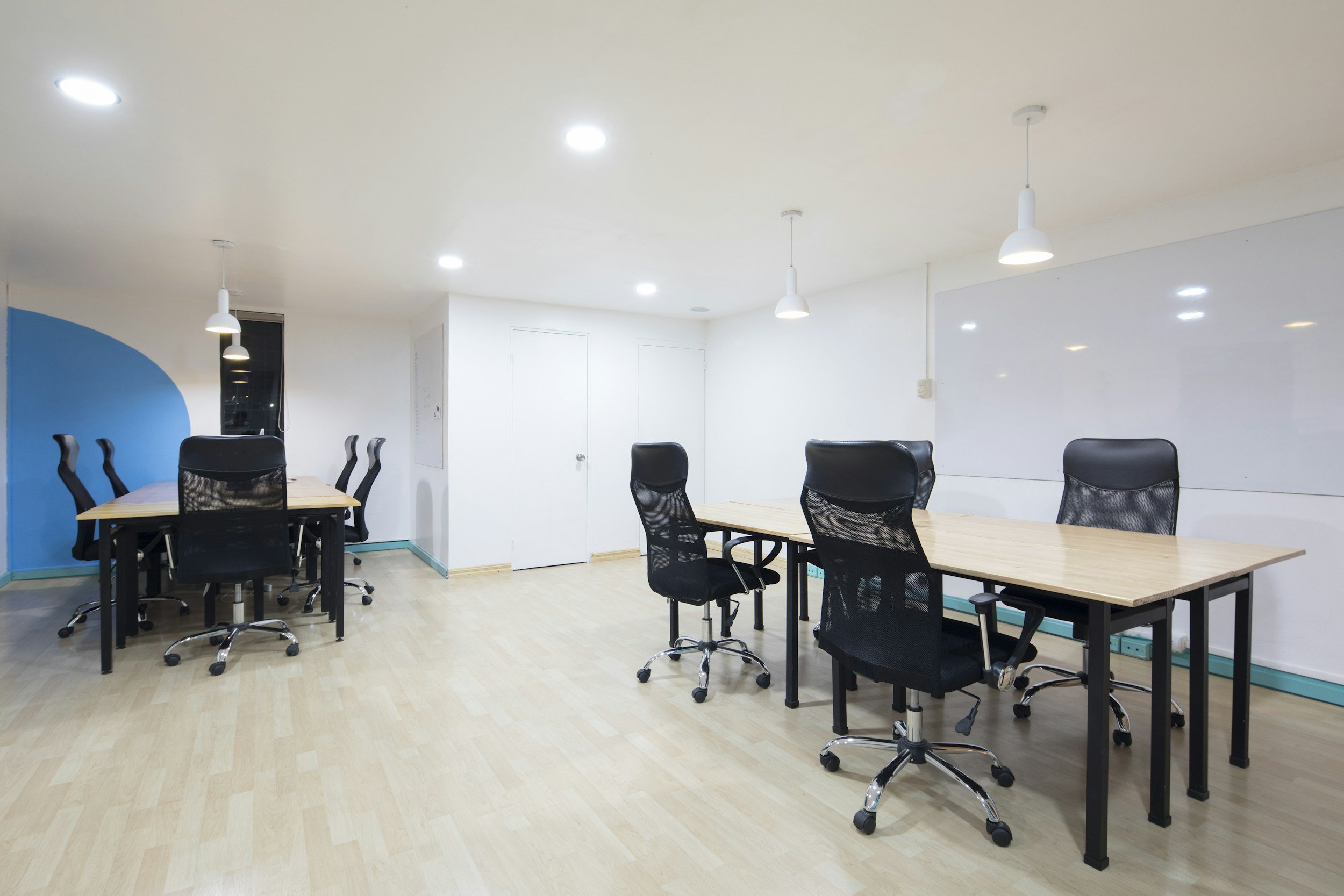 A modern conference room with multiple black office chairs around long light wood tables, white walls, ceiling-mounted lights, and a whiteboard on one wall.