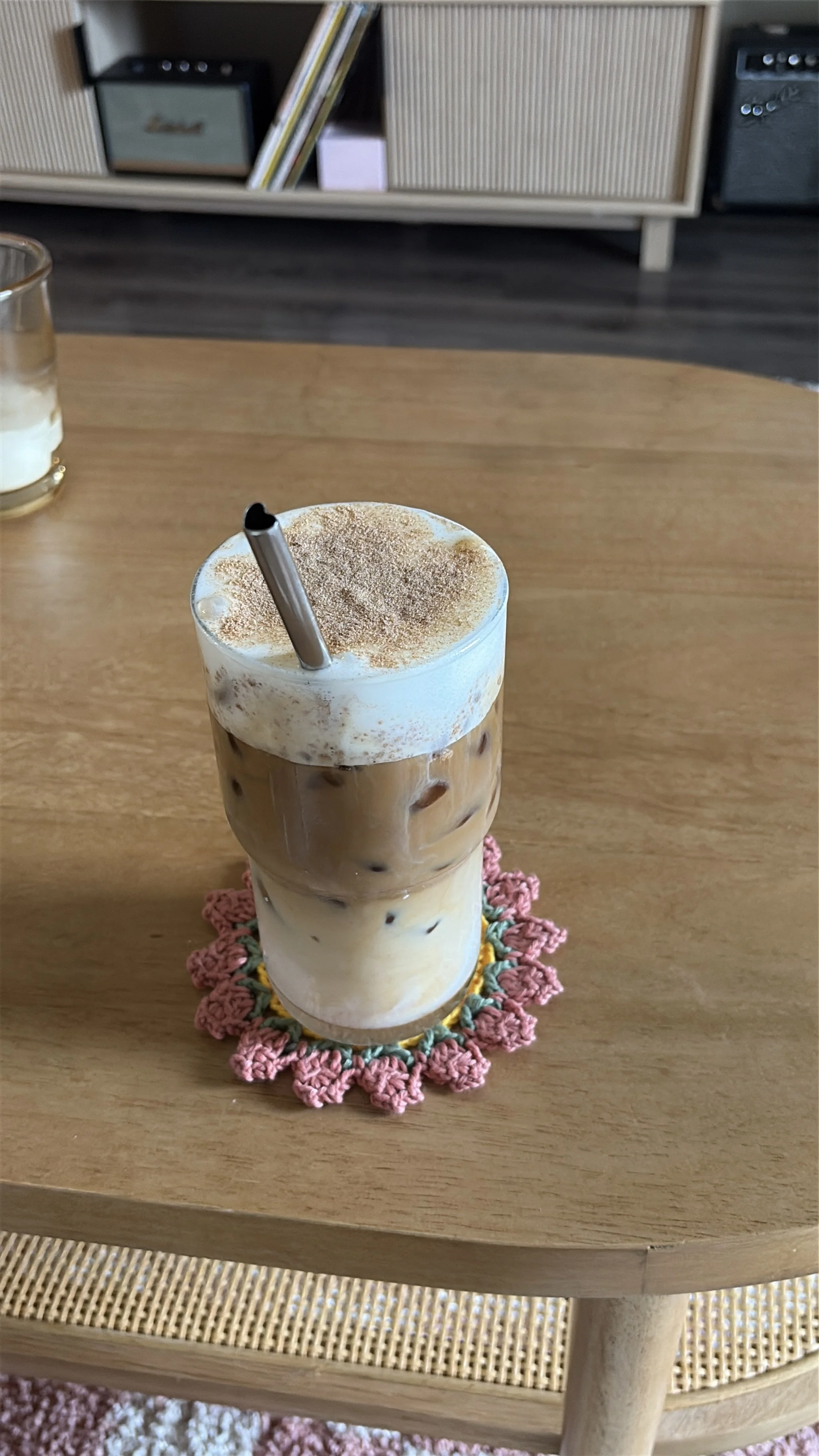 A layered iced coffee drink topped with whipped cream and cinnamon, served with a metal straw on a crocheted coaster on a wooden table.