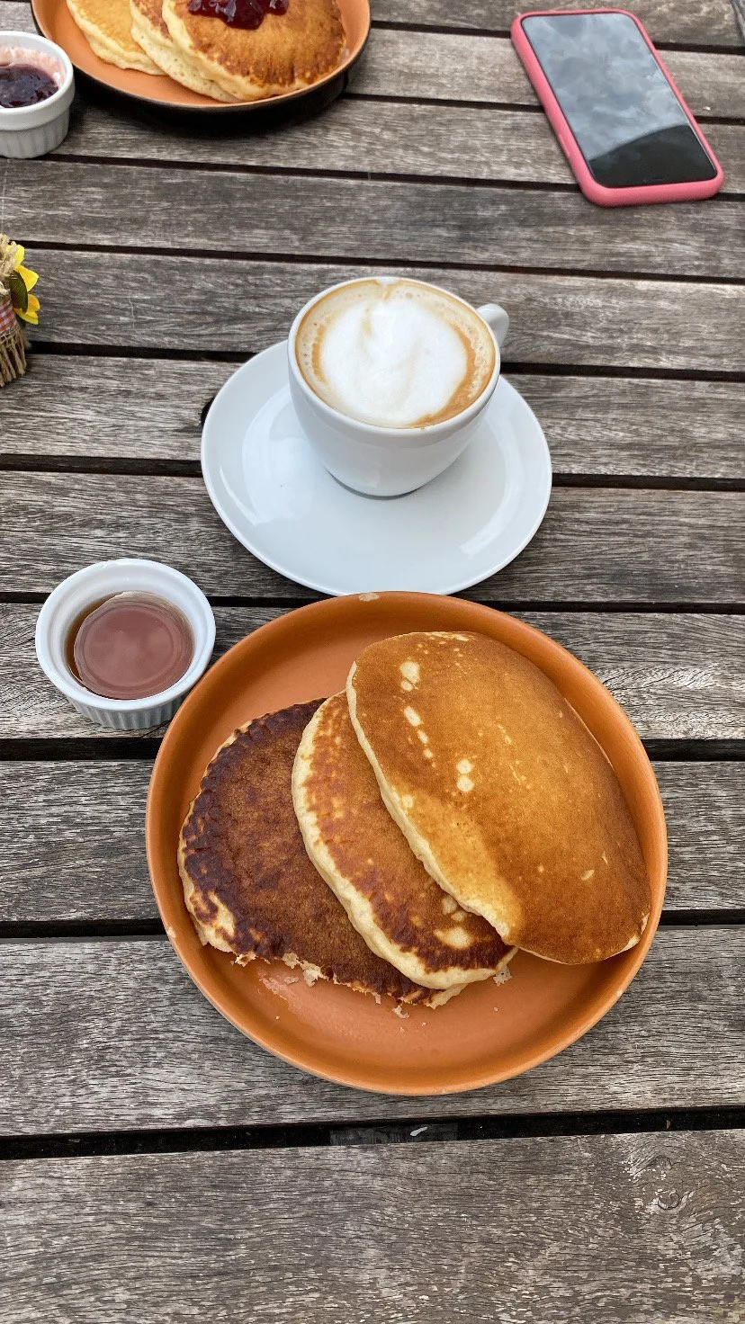 A wooden table with a plate of three pancakes, a cup of latte with foam, a small bowl of syrup, and another plate with additional pancakes. There is also a pink smartphone and a small dish of jam on the table.