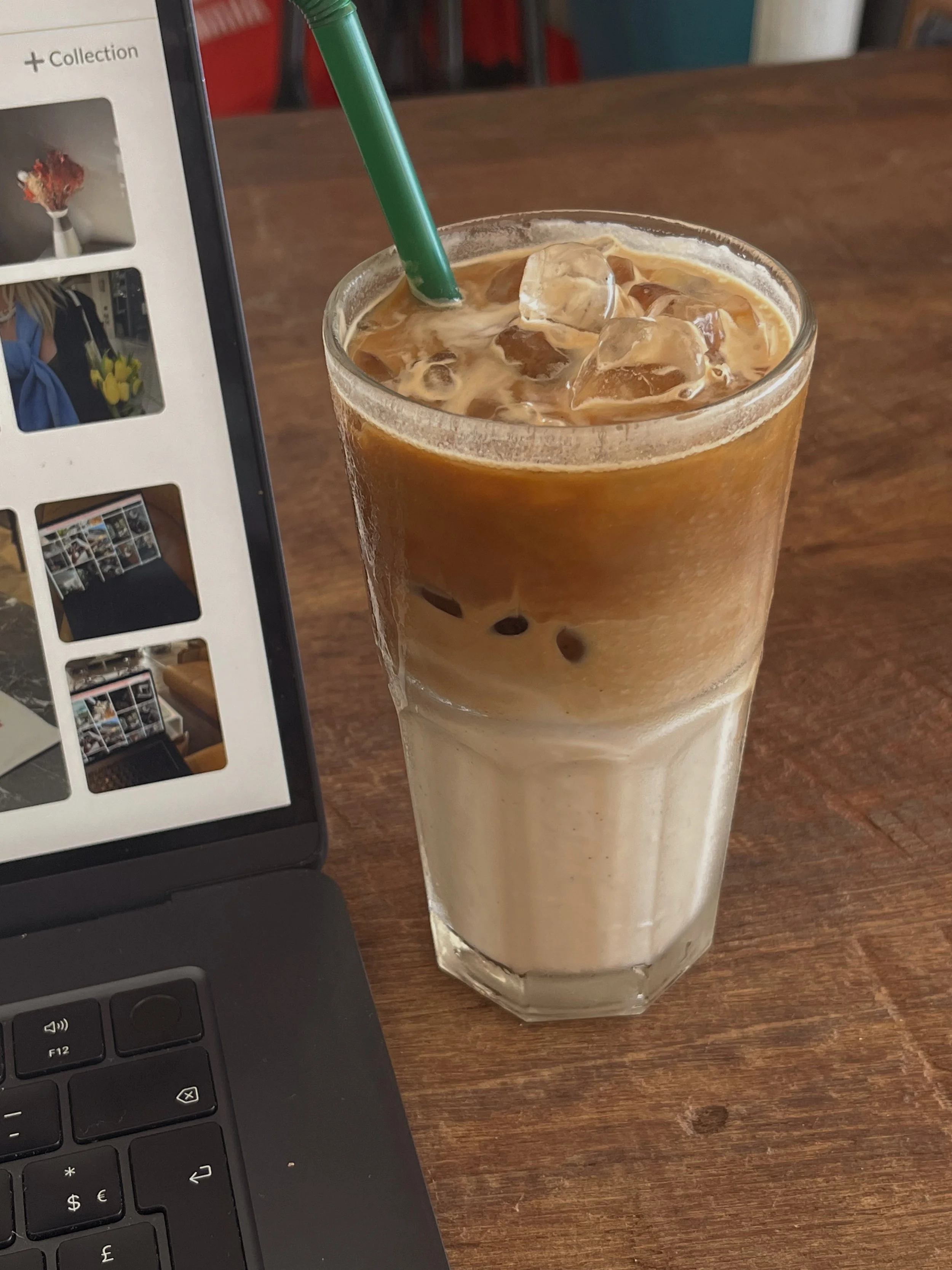 A tall glass of iced coffee with cream and ice cubes, with a green straw, placed on a wooden table next to a laptop.