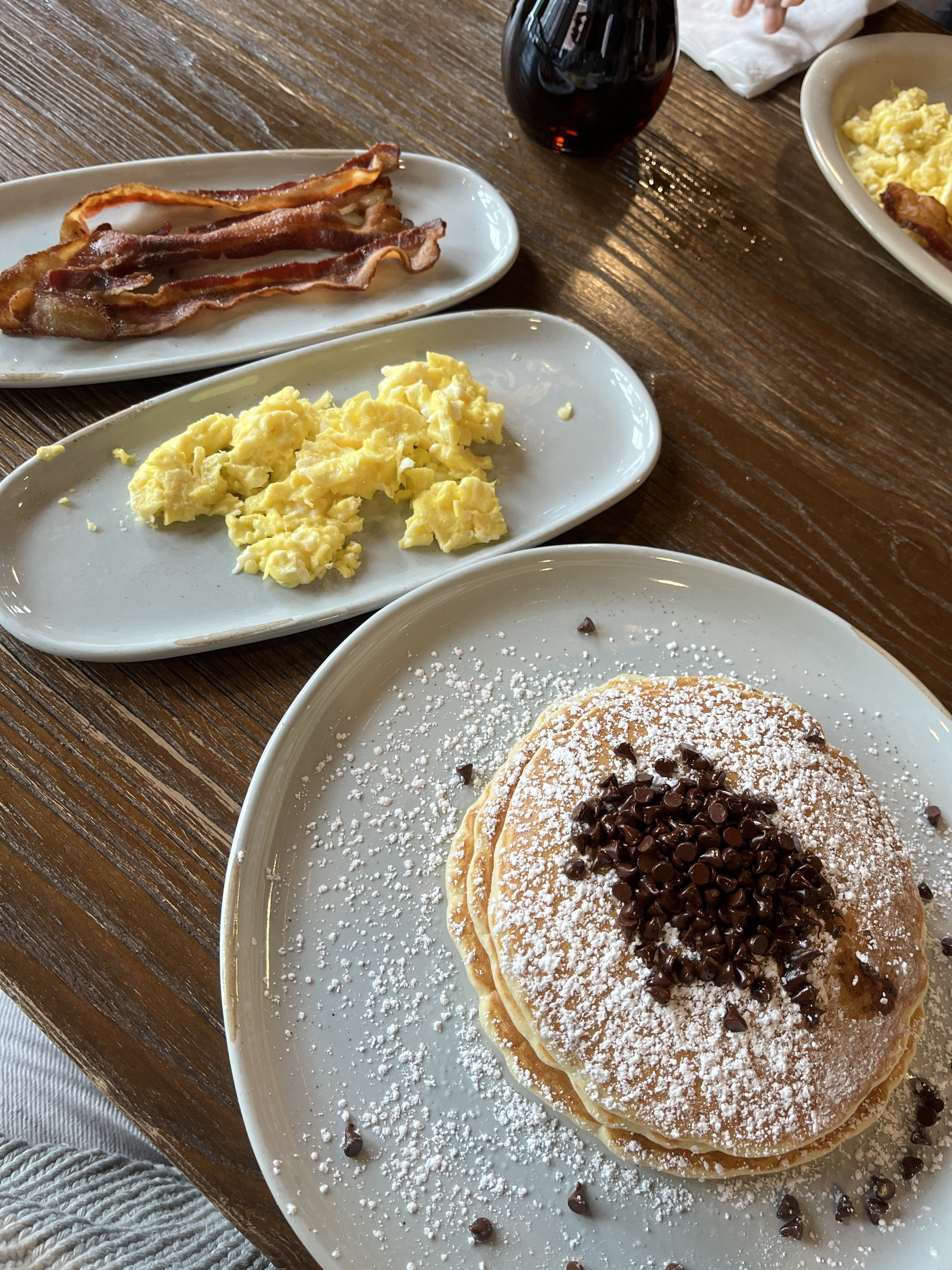 Breakfast consisting of chocolate chip pancakes with powdered sugar, scrambled eggs, and bacon on a wooden table.