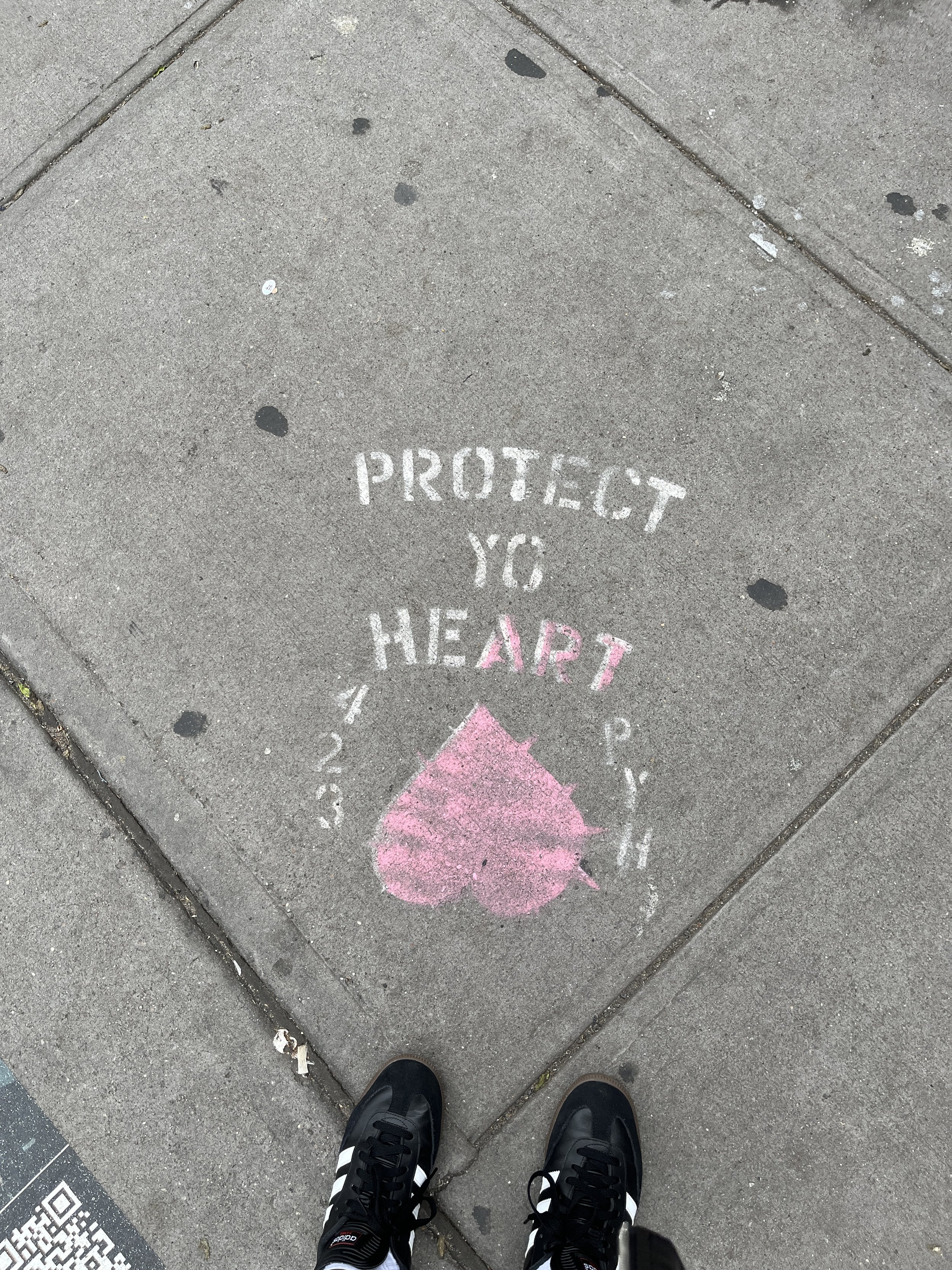 Sidewalk graffiti with the message 'Protect Yo Heart' in white and a pink heart design, with feet wearing black sneakers visible at the bottom.