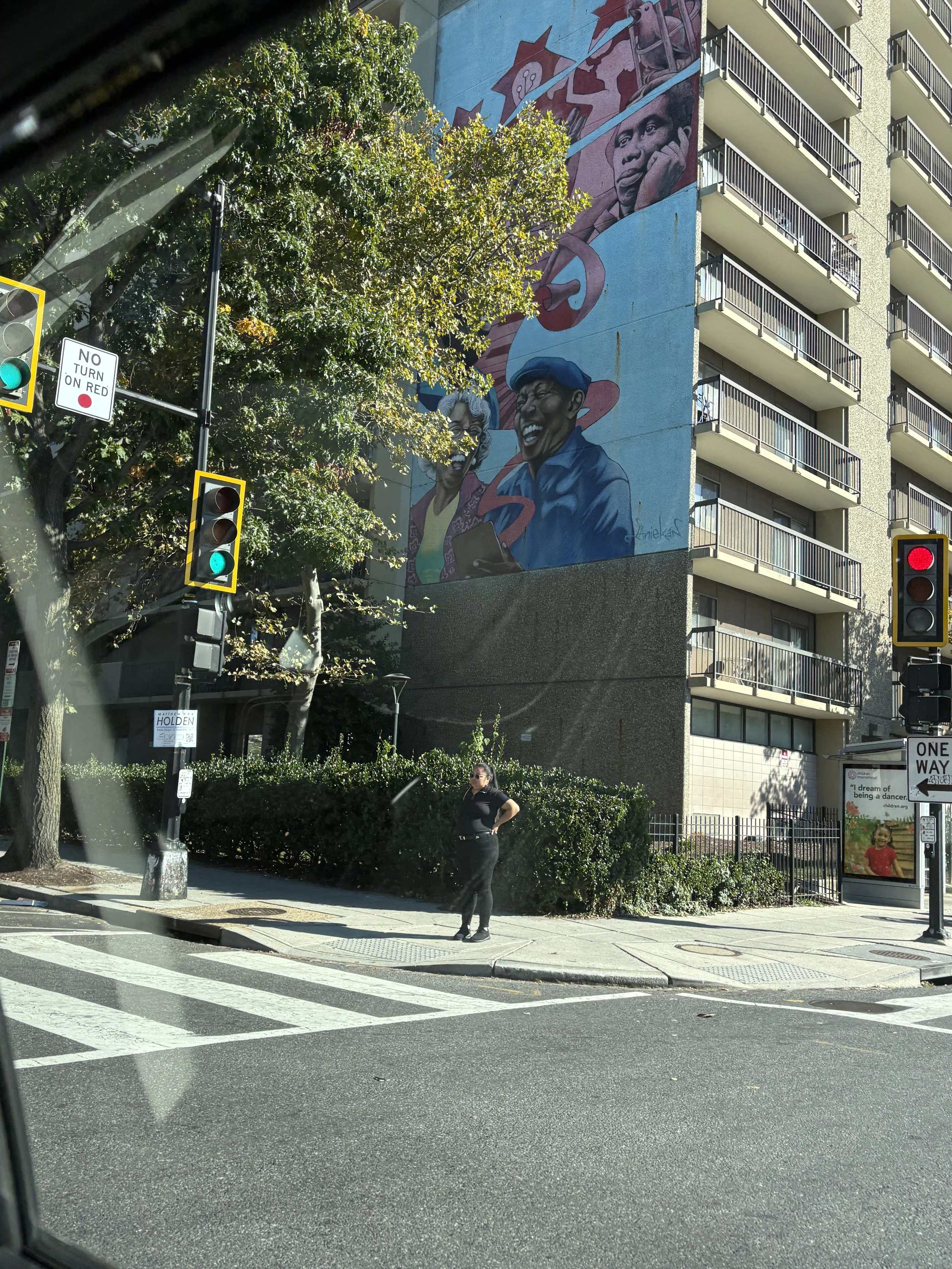 Street scene with traffic lights, a woman standing on the sidewalk, and a large mural on a building wall featuring caricatured portraits of people and vibrant colors.