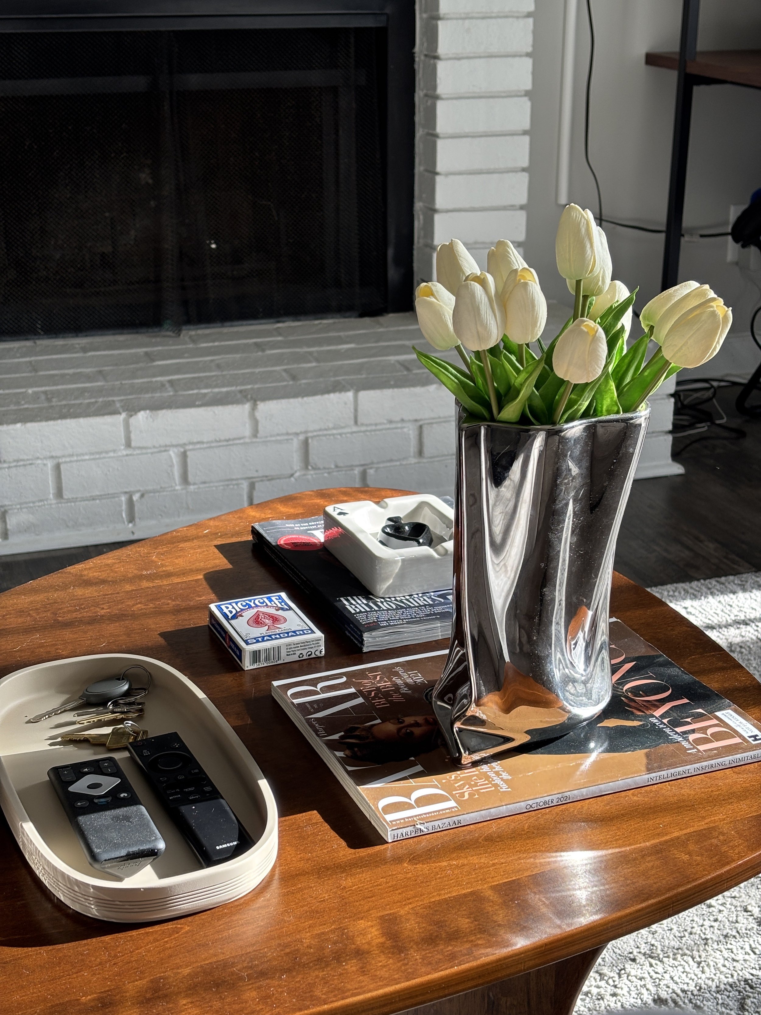 A living room scene with a coffee table featuring a metallic vase with white tulips, remote controls and keys in a tray, magazines, and a pack of playing cards, near a white brick fireplace.