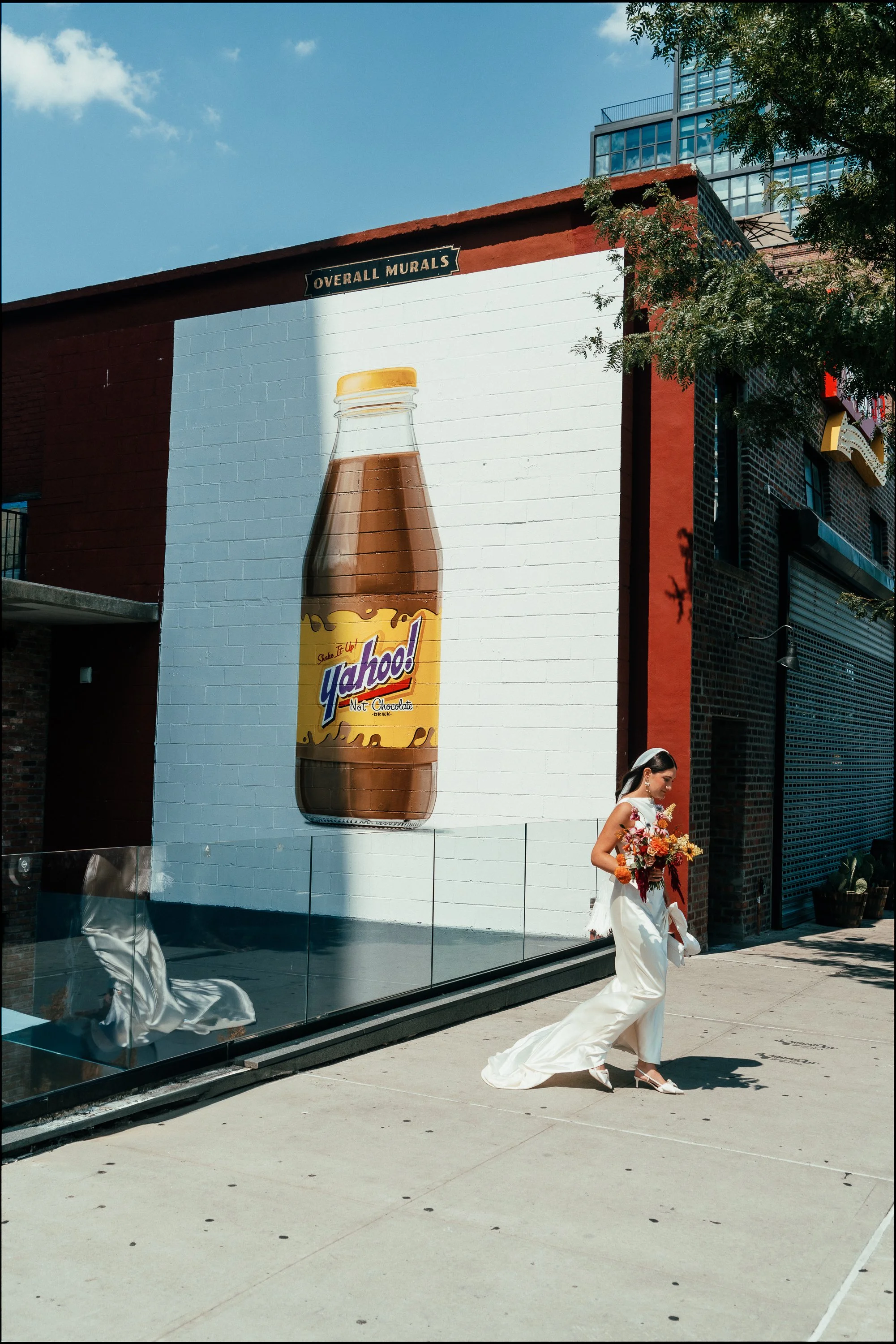 Street scene with a large wall mural of a Yoo-hoo chocolate drink bottle and a woman in wedding attire holding a bouquet walking past.