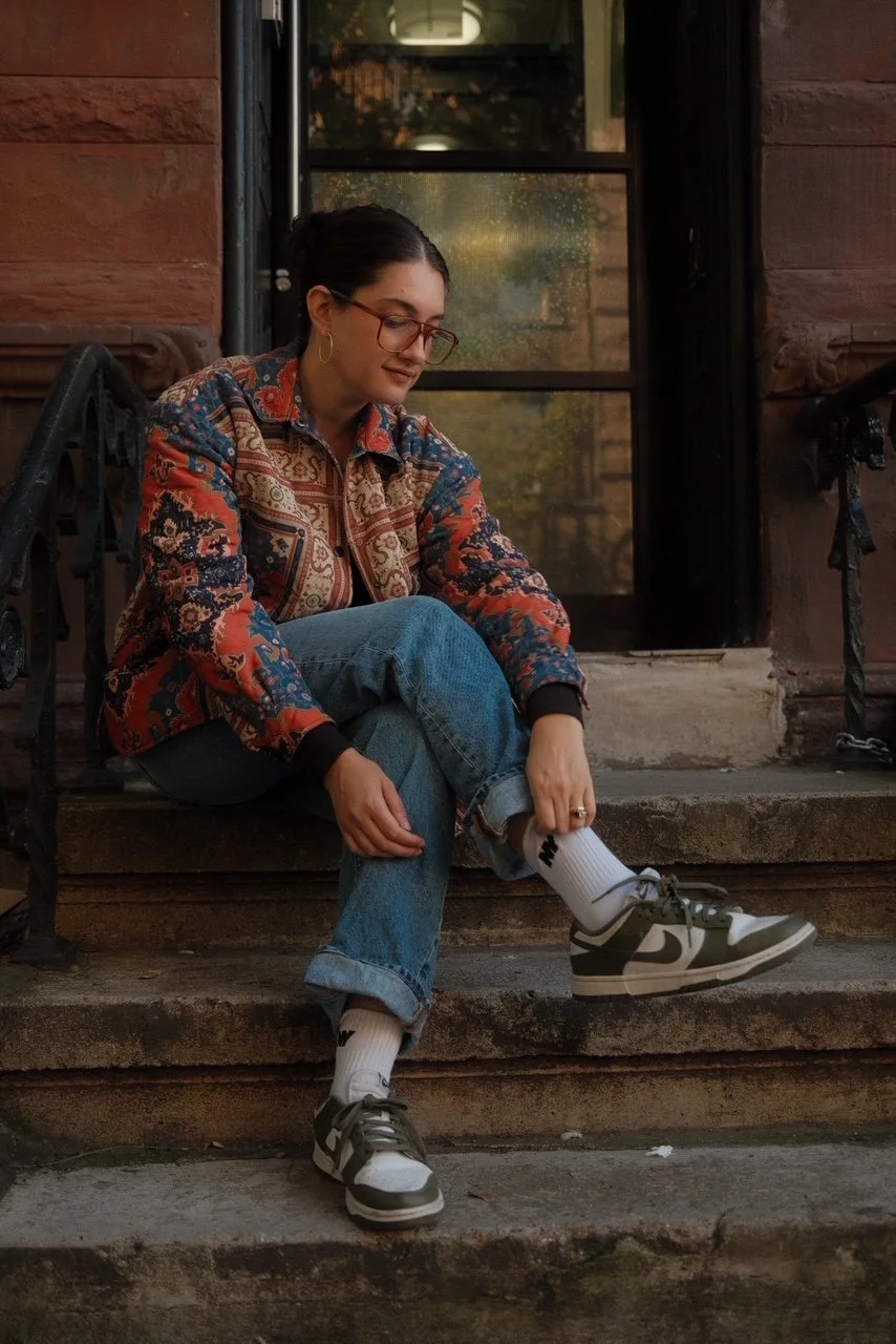 A woman sitting on outdoor stairs, wearing glasses, a colorful patterned shirt, jeans, white socks, and Nike sneakers, adjusting her sock.