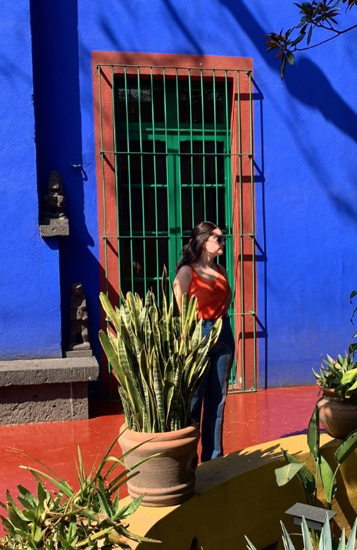 A woman with dark hair in an orange sleeveless top stands outside in front of a vibrant blue wall with a green door, surrounded by potted plants.
