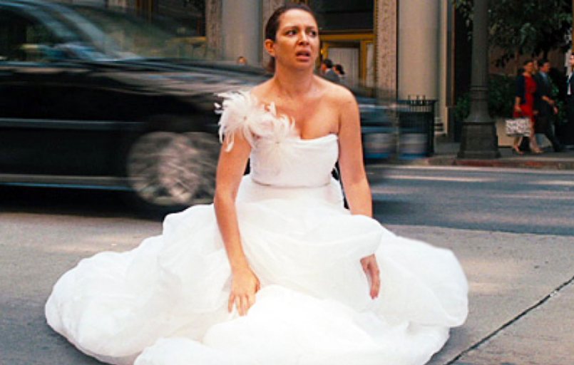 A woman wearing a white dress is kneeling on the street, looking distressed, with a blurred car passing in the background.