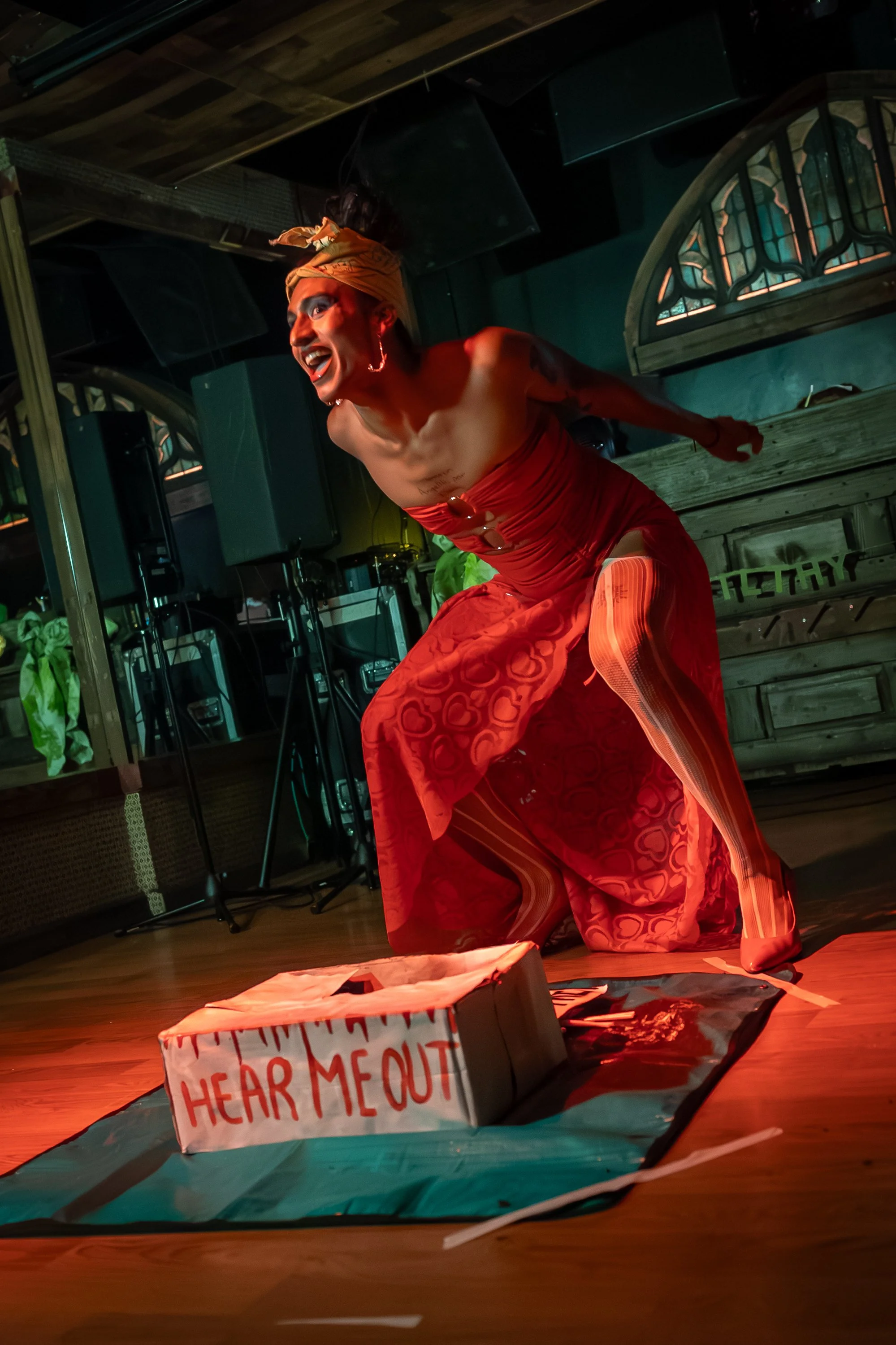 Performer with makeup and a headwrap on a stage, wearing a red dress, leaning forward with an intense expression, surrounded by music equipment and a cardboard box with 'HEAR ME OUT' written on it.