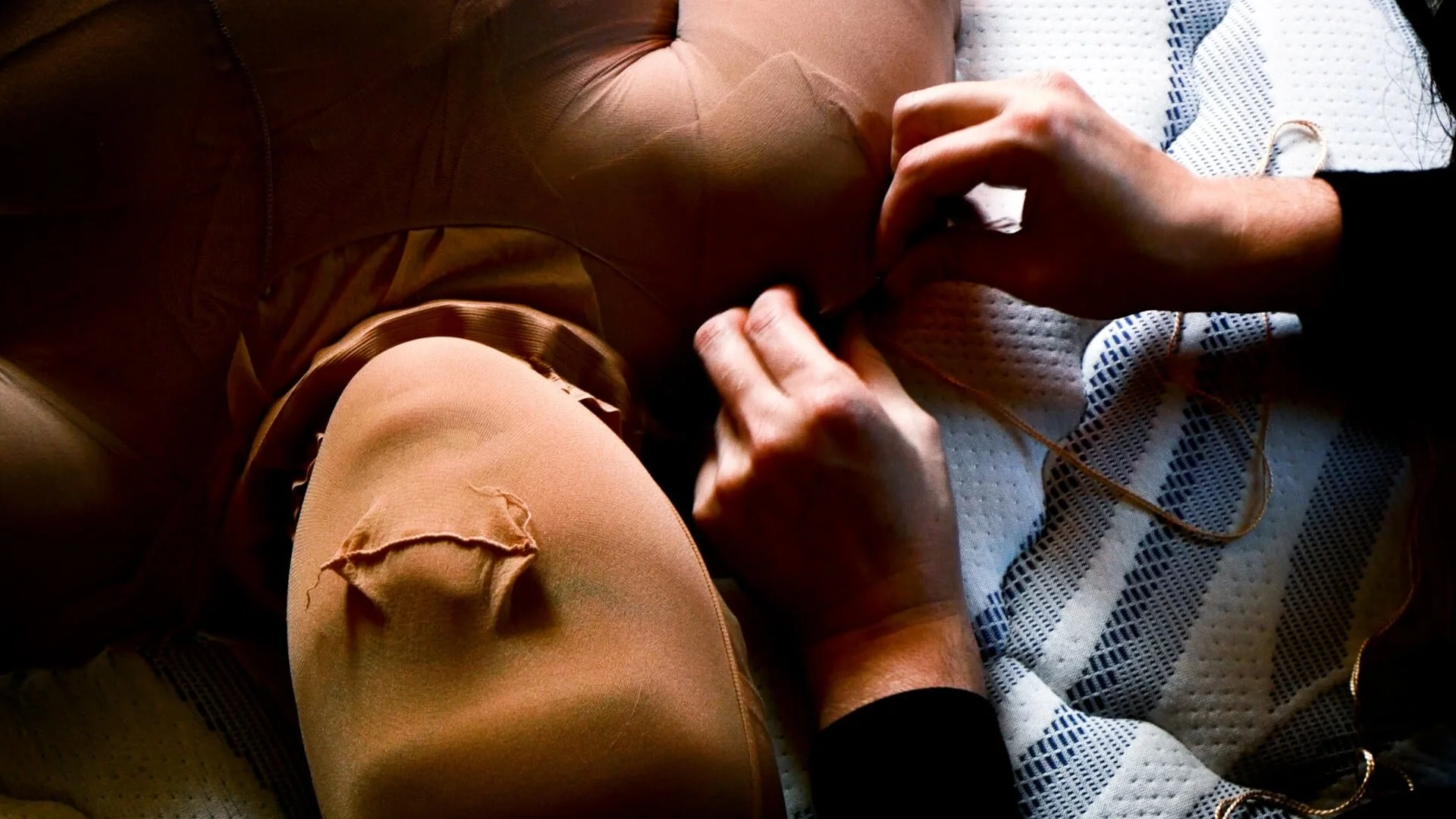 Close-up of a person sewing or repairing a brown leather item, possibly a bag, with their hands using a needle and thread on a white and blue checkered fabric surface.