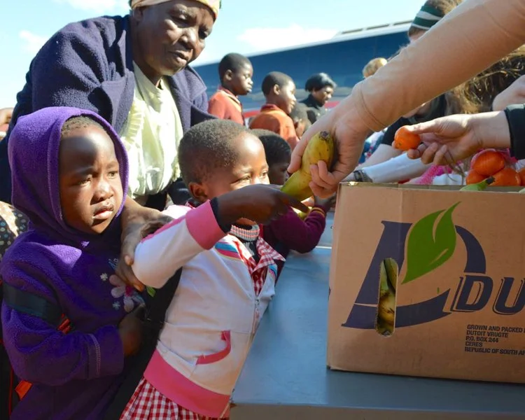 Children in line receiving vegetables from volunteers at a food distribution event, with a man reaching out to give a banana and others holding carrots, in an outdoor setting.