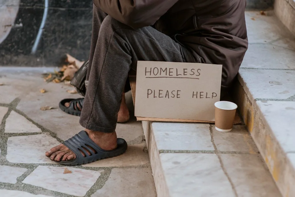 A homeless person sitting on a sidewalk with a handmade sign that reads 'HOMELESS PLEASE HELP' and a paper cup for donations.