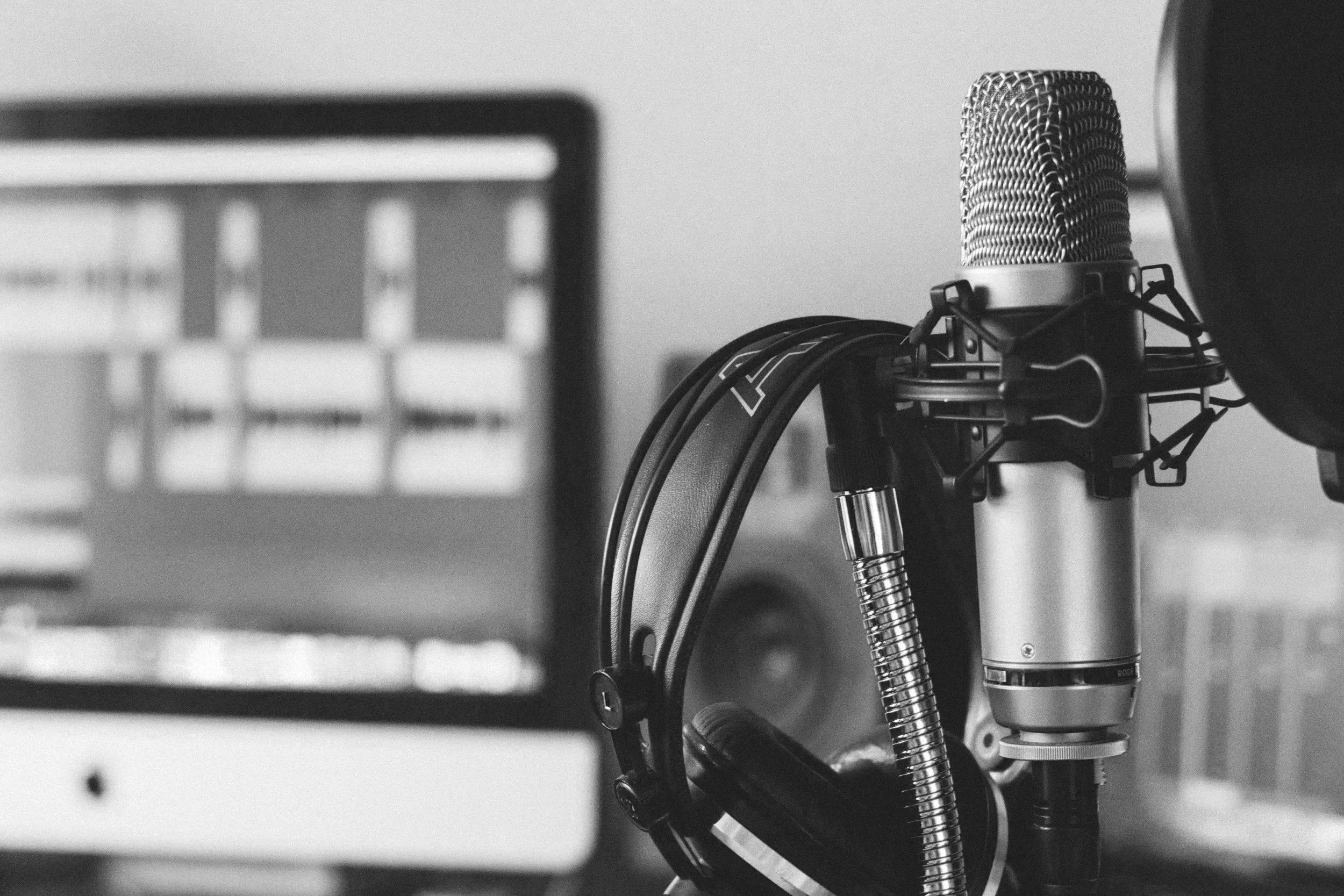 Close-up of a professional studio microphone with headphones hanging on a table, blurred computer monitor in the background.