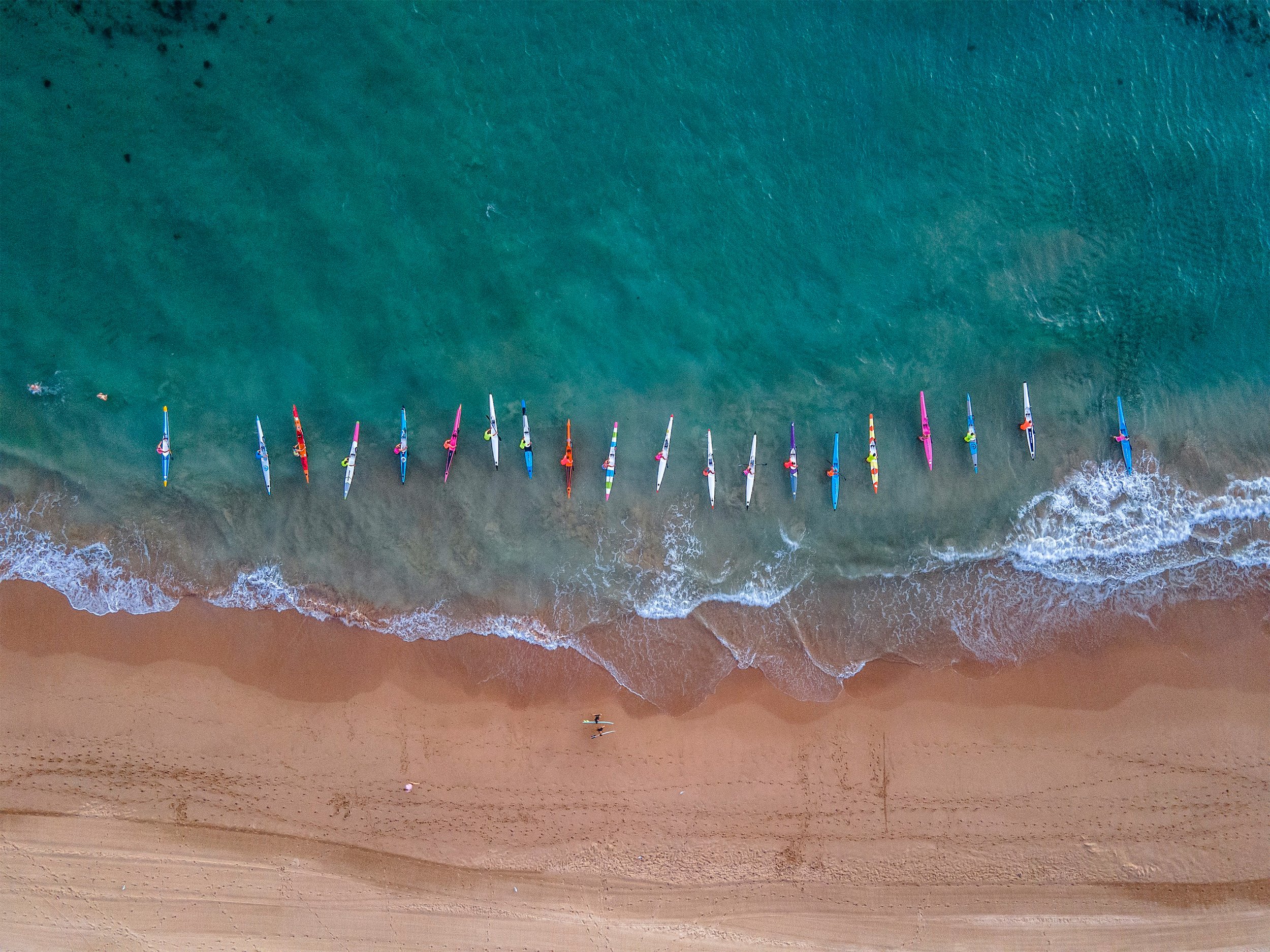 Aerial view of colorful sailboats lined up on a sandy beach, with two people standing near the water and a person paddleboarding in the ocean.