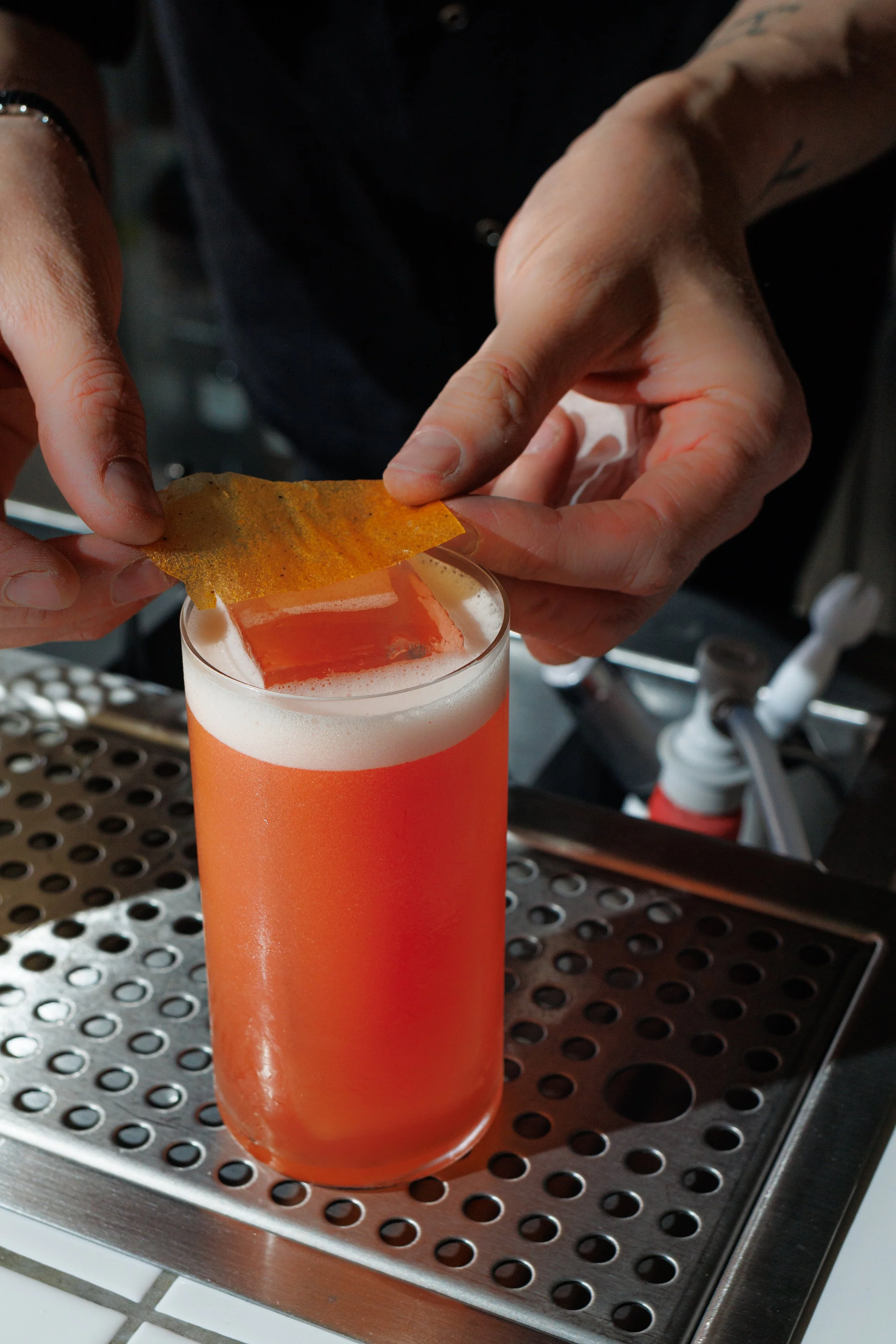 A bartender is placing a gold-colored garnish onto a bright orange cocktail in a tall glass.