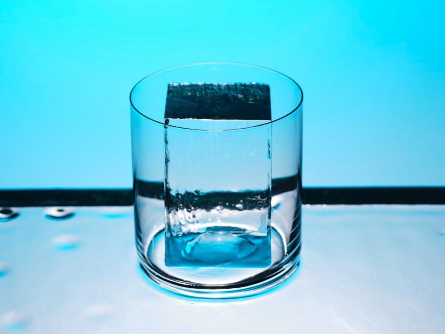 A glass with a large ice cube inside it, against a blue background.