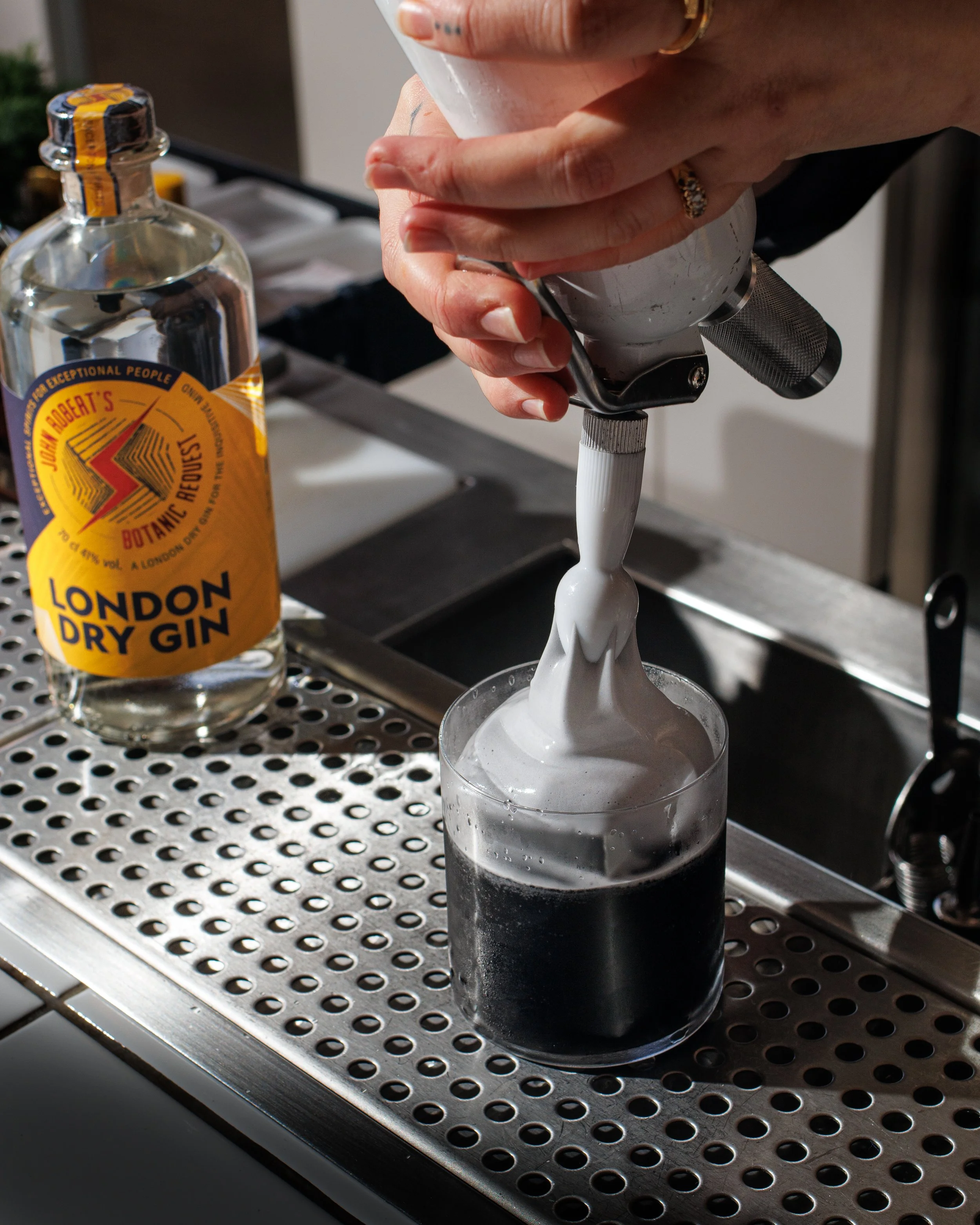 A bartender uses a siphon to dispense foam onto a dark cocktail in a clear glass, with a bottle of London dry gin on a metal bar counter nearby.