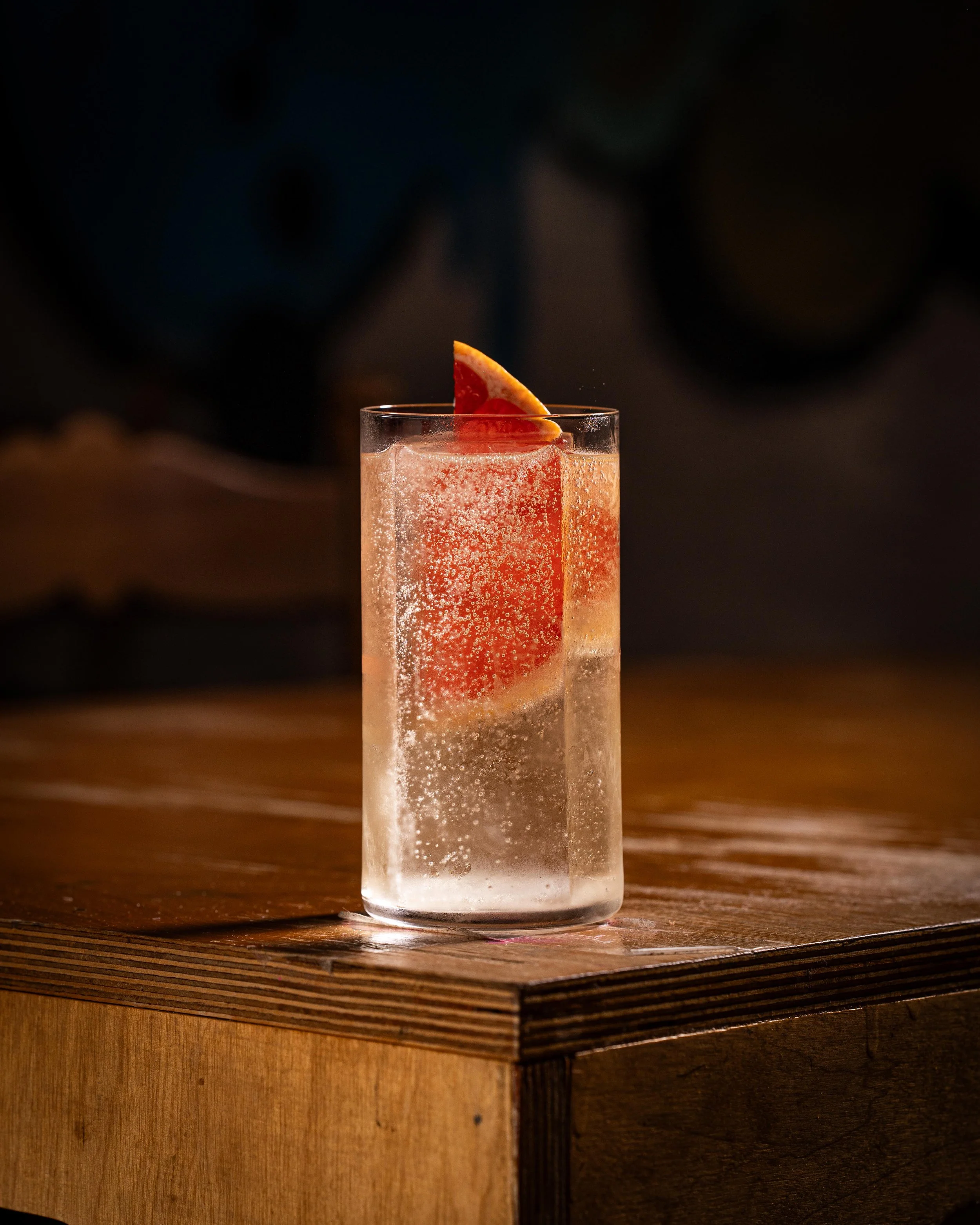 A clear glass filled with a sparkling, fizzy pink drink, garnished with a wedge of grapefruit, sitting on a wooden surface.