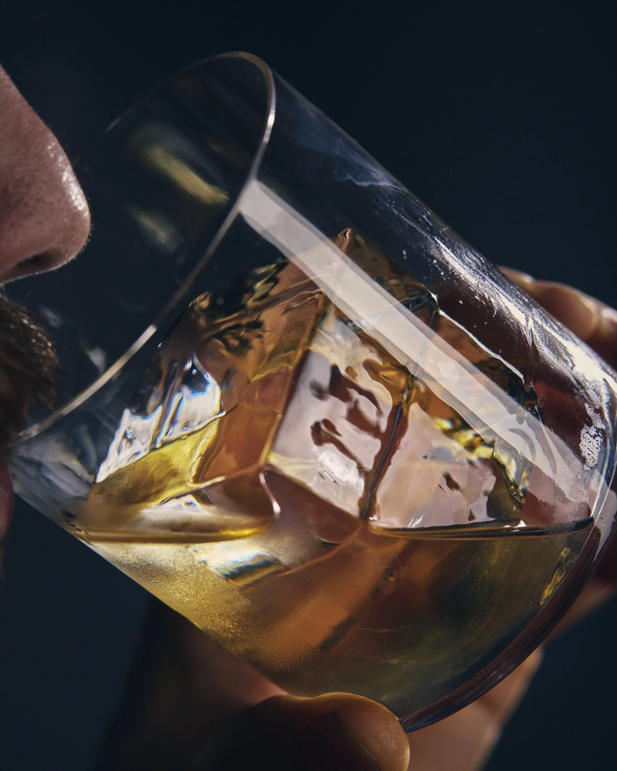 Close-up of a person drinking a glass of amber-colored wine with ice, against a dark background.