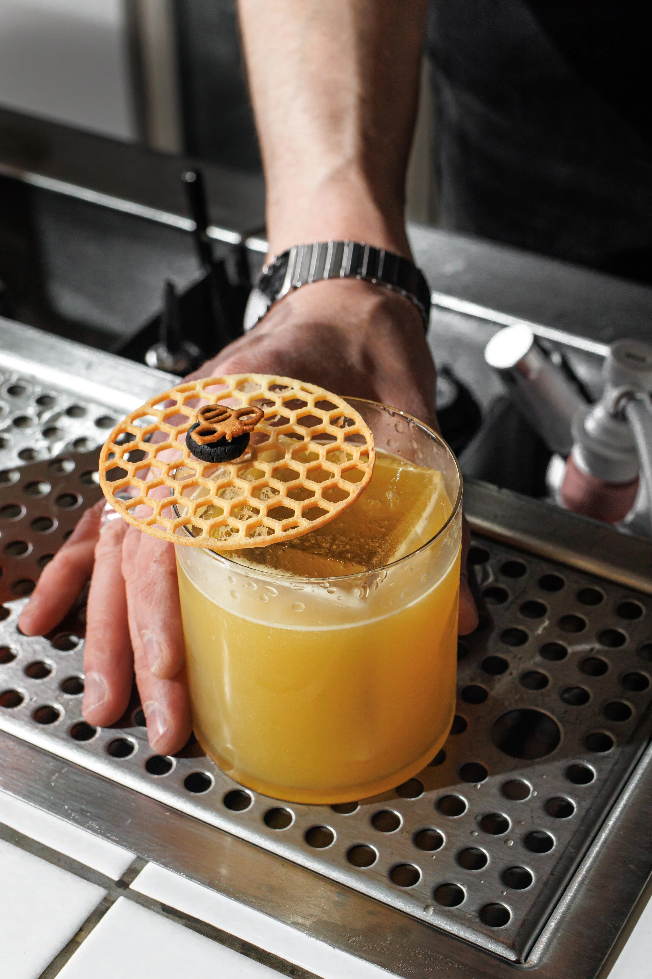 A bartender's hand resting on a metallic bar counter, with a yellow cocktail in front garnished with a crispy honeycomb-patterned wafer adorned with a small decorative element.