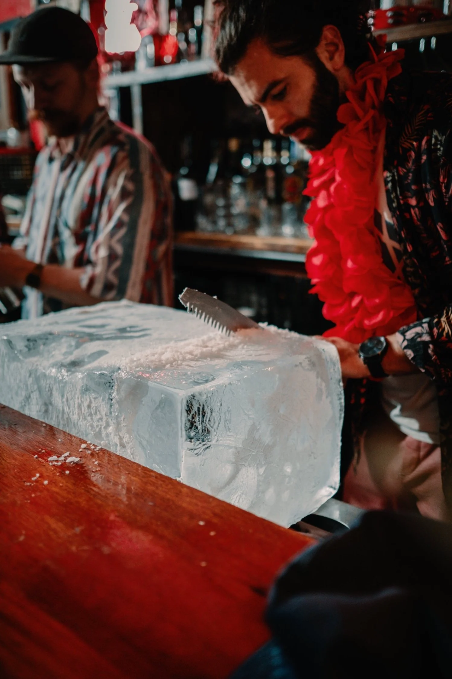 A man wearing a red lei and floral shirt is carving a block of ice with a saw at a bar. There is a woman in a striped shirt in the background.