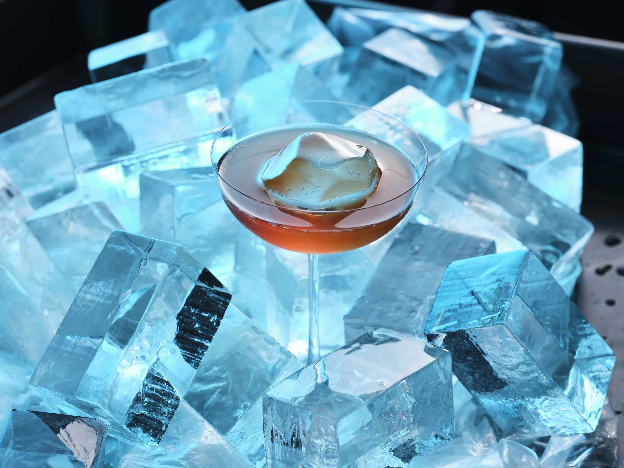 A cocktail in a coupe glass with a large ice cube, surrounded by ice cubes with a blue light glow.