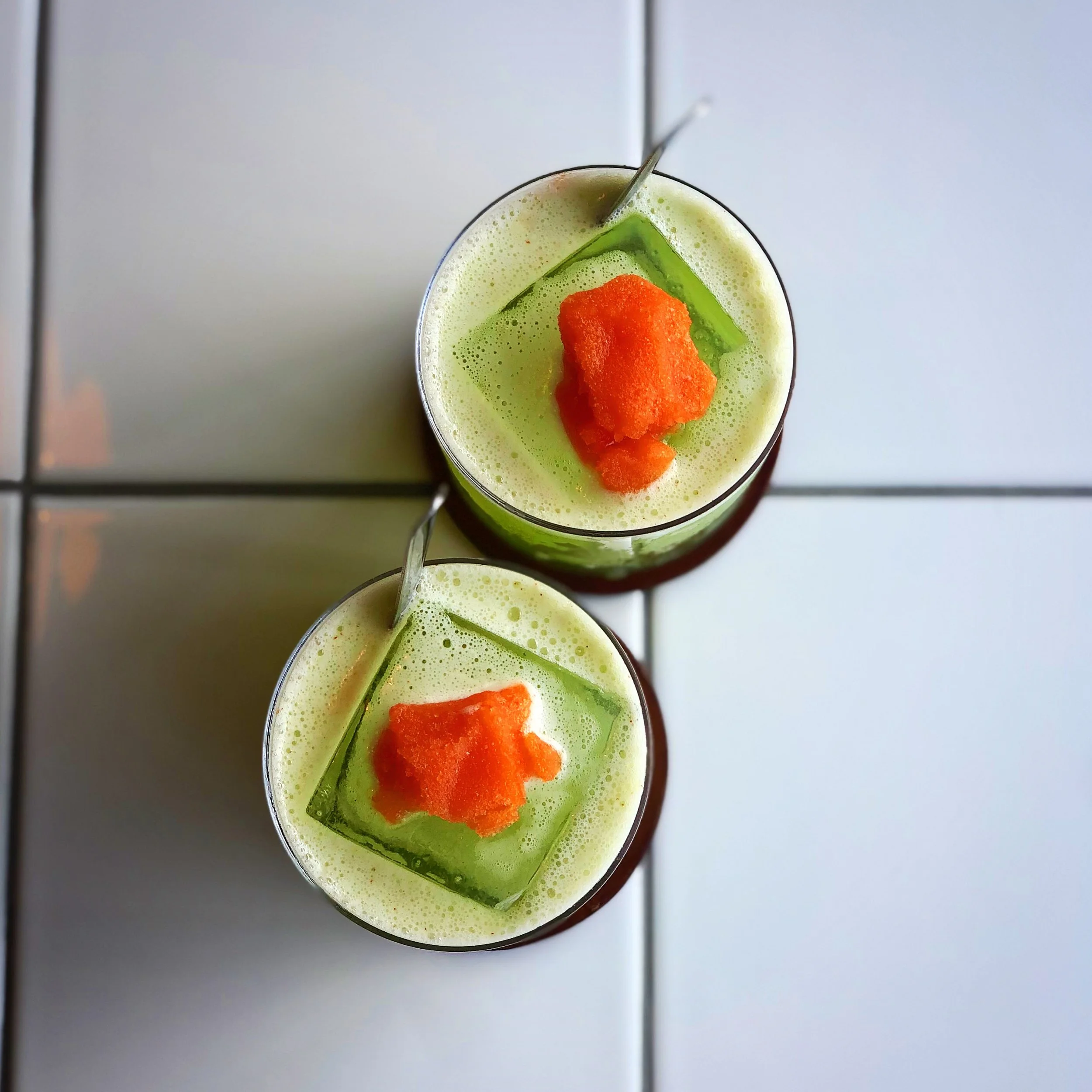 Two glasses of green frothy beverage topped with orange-colored puree and served with a green ice cube on a white tiled surface.