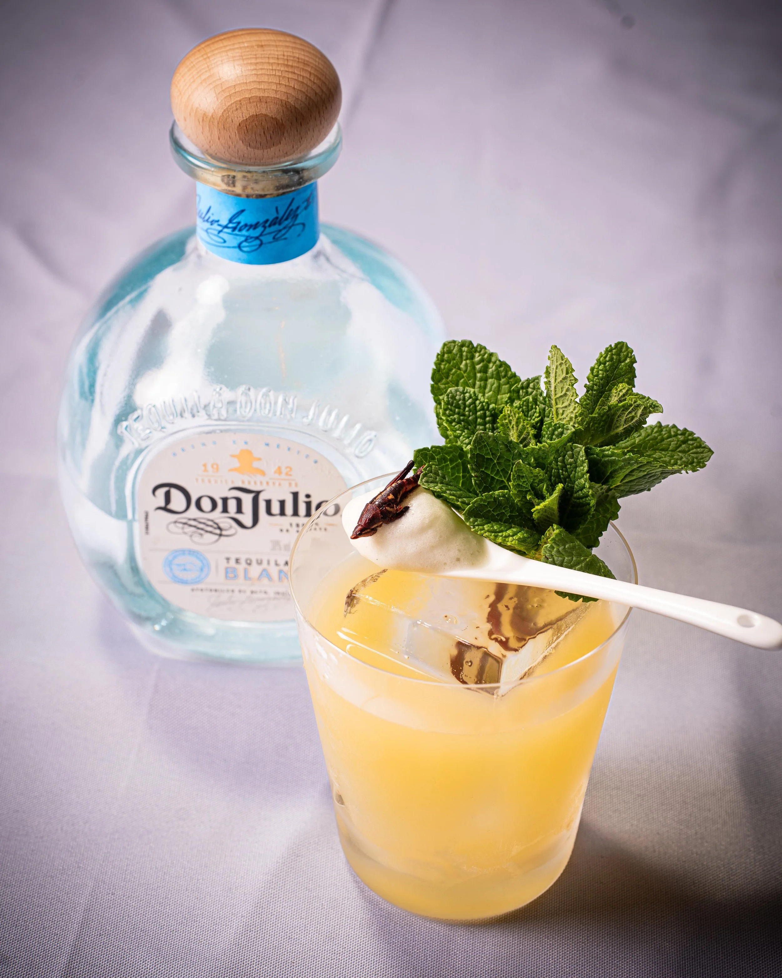 A cocktail garnished with fresh mint leaves, a sprig of rosemary, and an ice cube with a decorative stir stick, next to a bottle of Don Julio Blanco tequila.