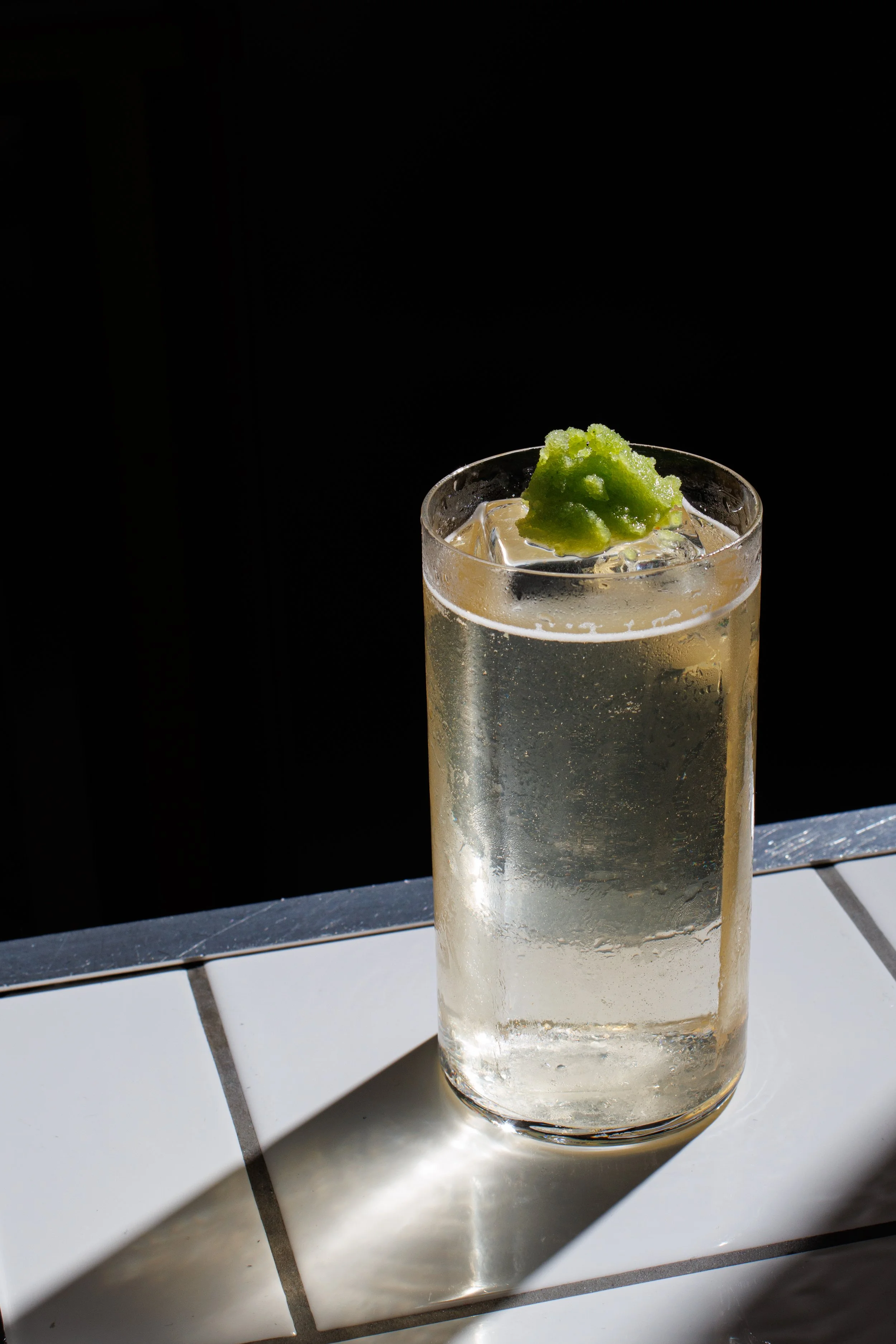 A glass of clear soda with ice cubes and a dollop of green wasabi on top, placed on a tiled surface with sunlight casting a shadow.