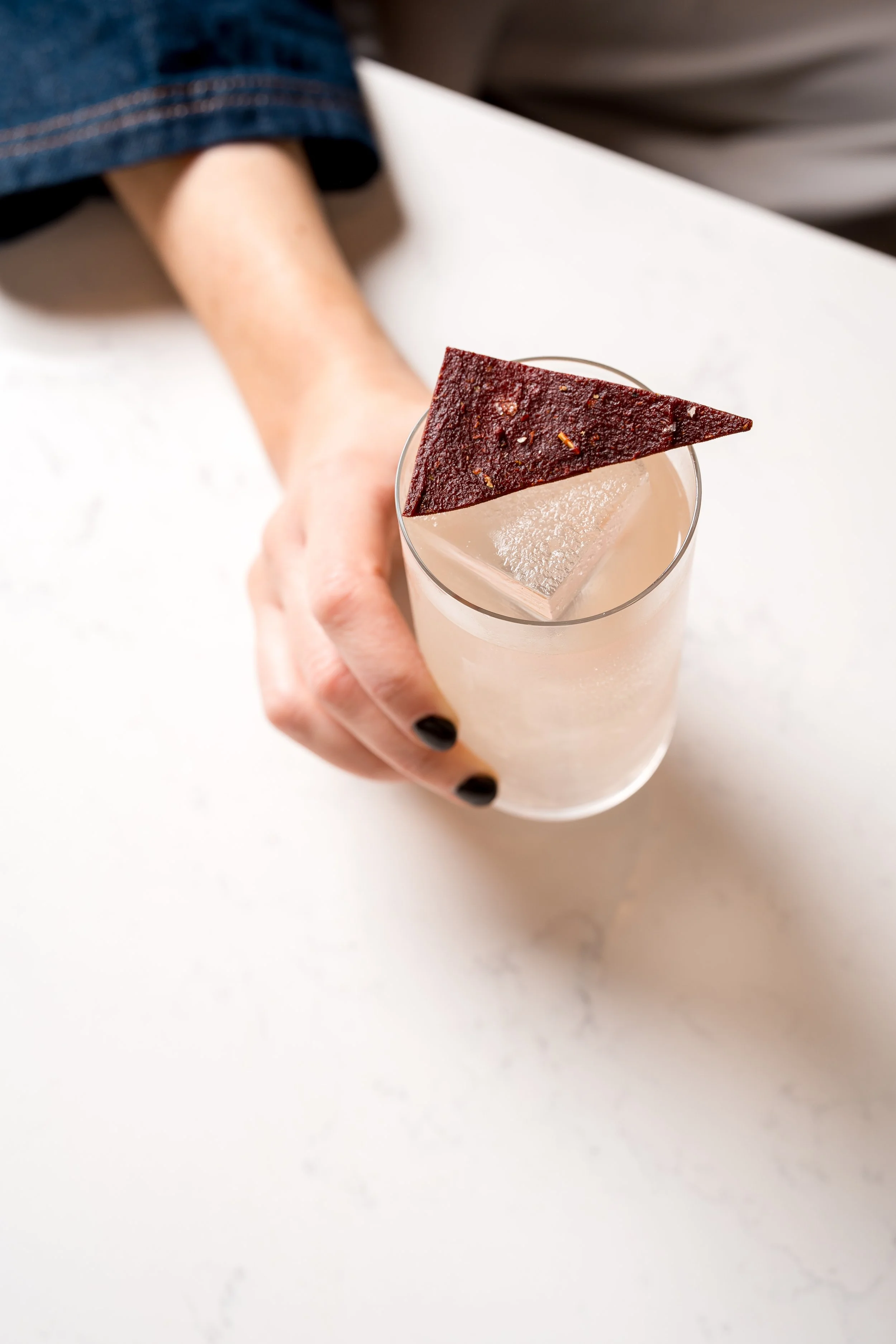 Person holding a clear glass of cocktail with a large ice cube and garnished with a triangular piece of dark red fruit leather.