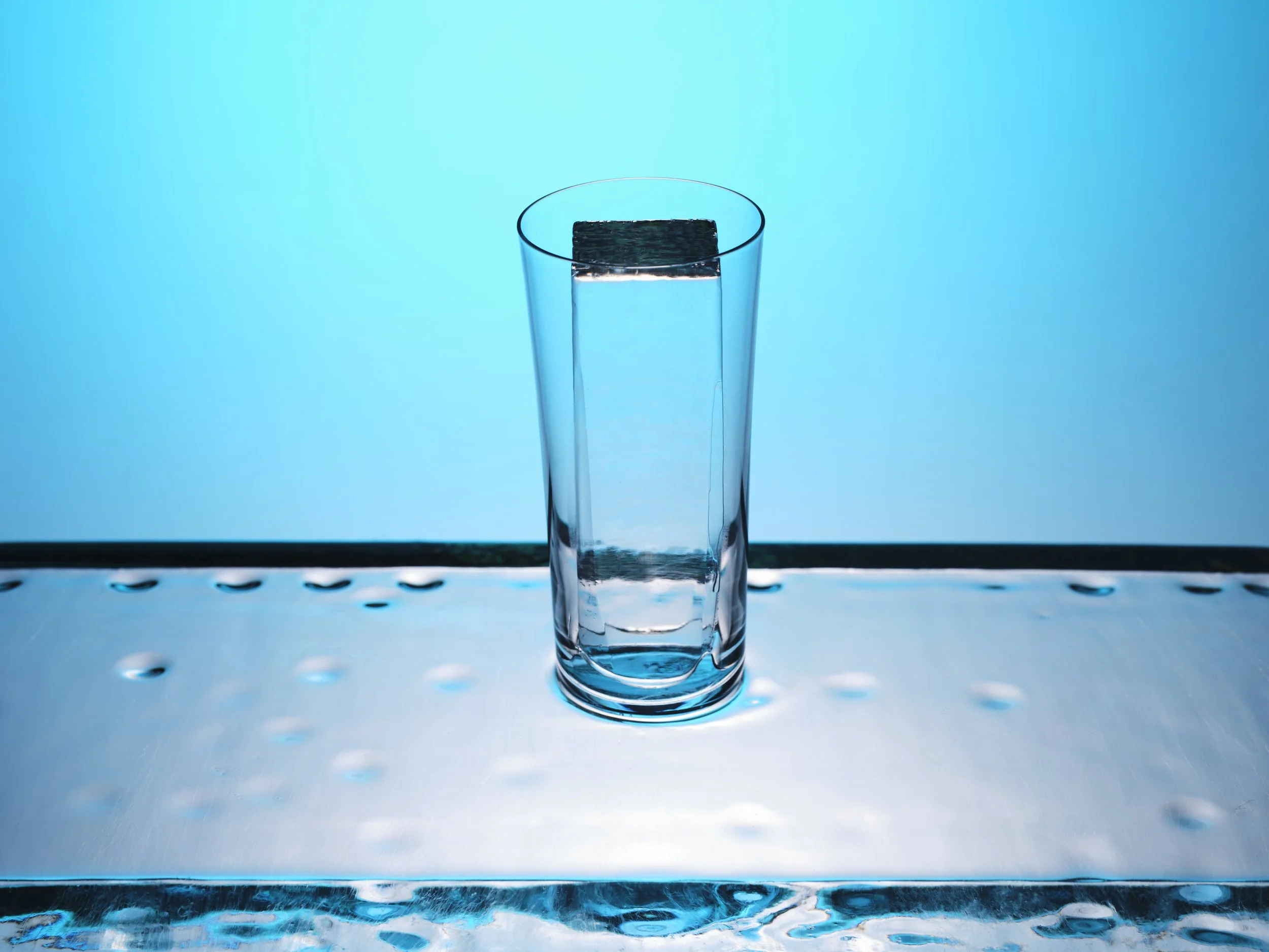 Tall clear glass filled with water, placed on a reflective surface with a blue background.