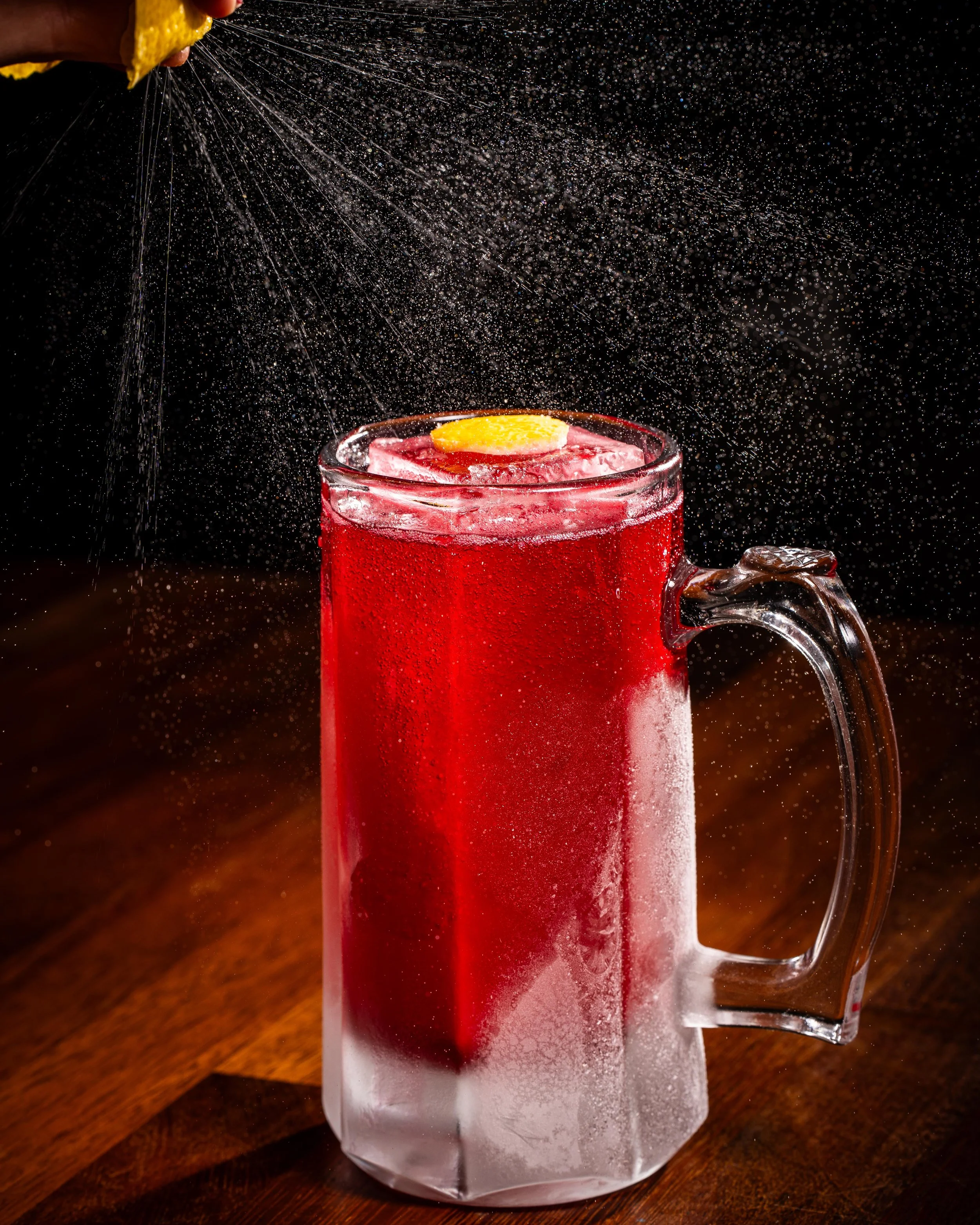 A glass mug of red iced beverage garnished with a lemon slice, with sparkling water being poured into it, creating bubbles and mist.