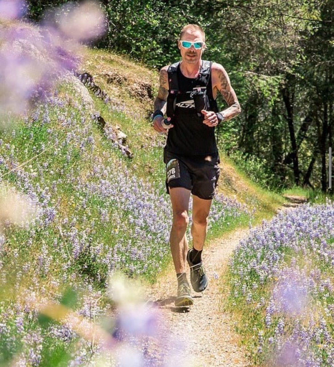 Trail runner in sunglasses and black athletic gear running along a narrow path lined with purple wildflowers, surrounded by a lush green forest.