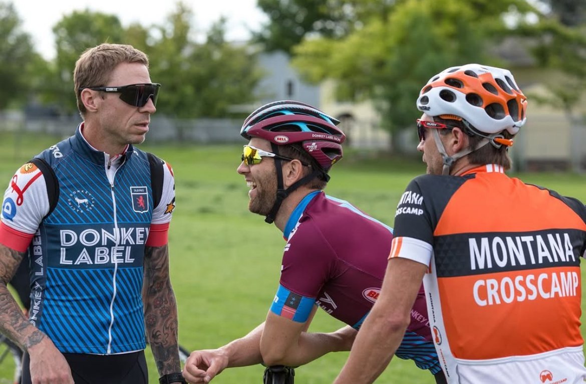 Three cyclists in conversation, wearing colorful cycling jerseys and helmets, standing on grass with trees in the background.