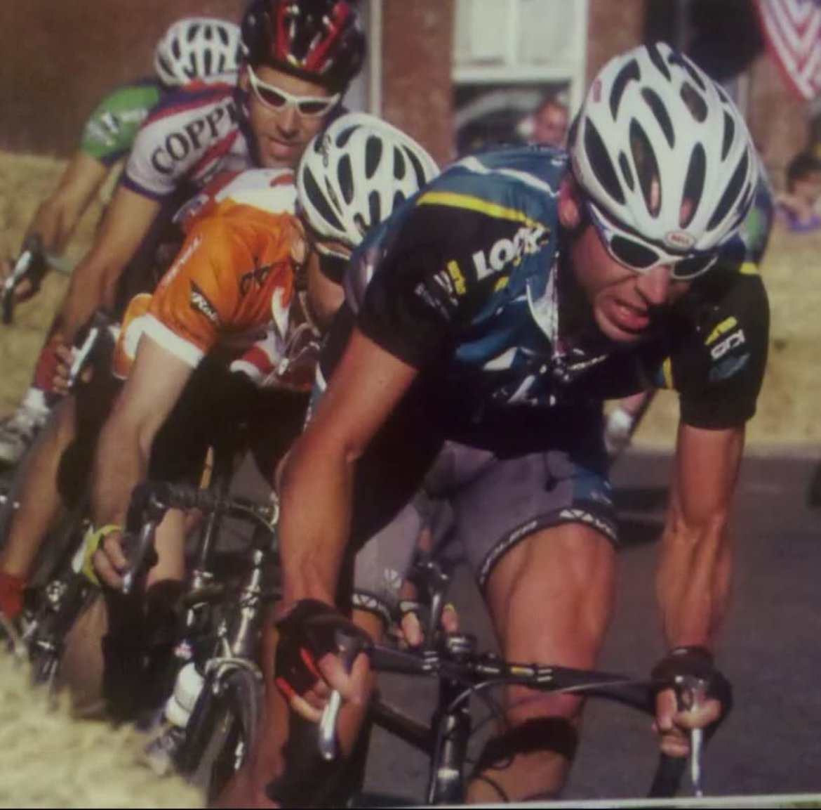 Cyclists in a road race, focused on speed and intensity. They wear helmets and cycling gear, navigating through a corner, with a blurred background suggesting motion.