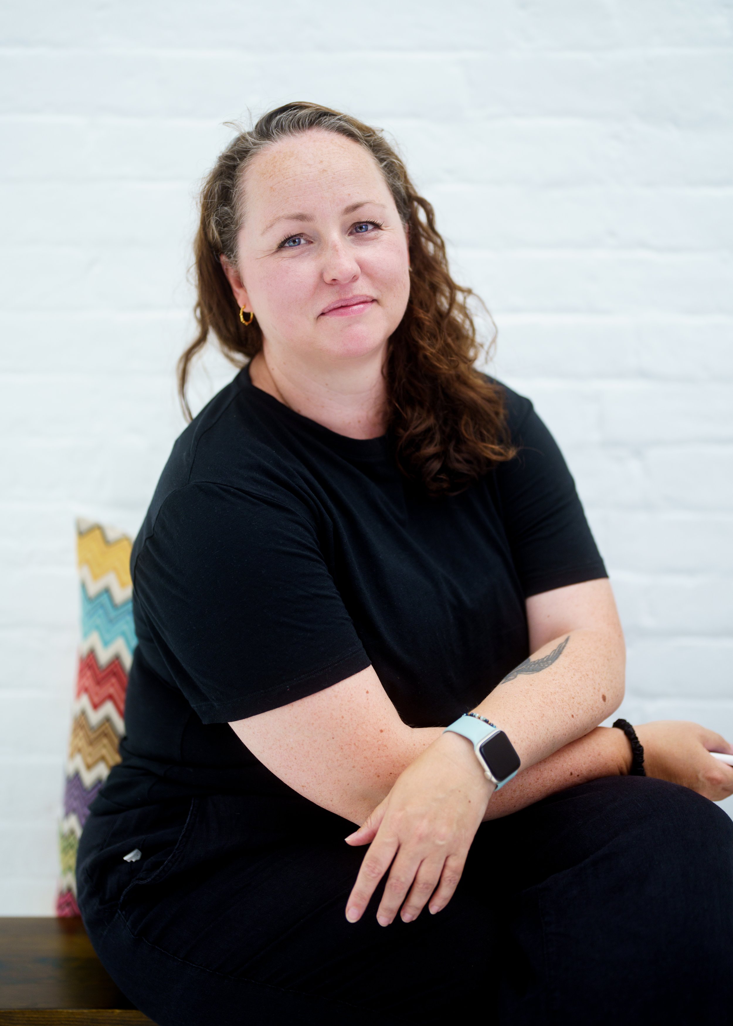 A woman with curly brown hair, wearing a black t-shirt, sitting with arms crossed in front of a white brick wall background.