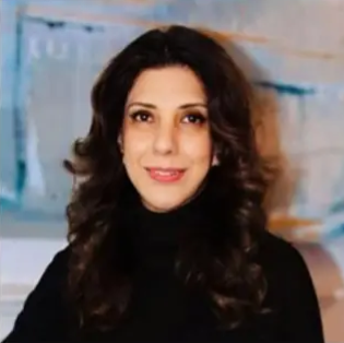 Portrait of a woman with dark, curly hair wearing a black top, smiling indoors with abstract blue and beige background.