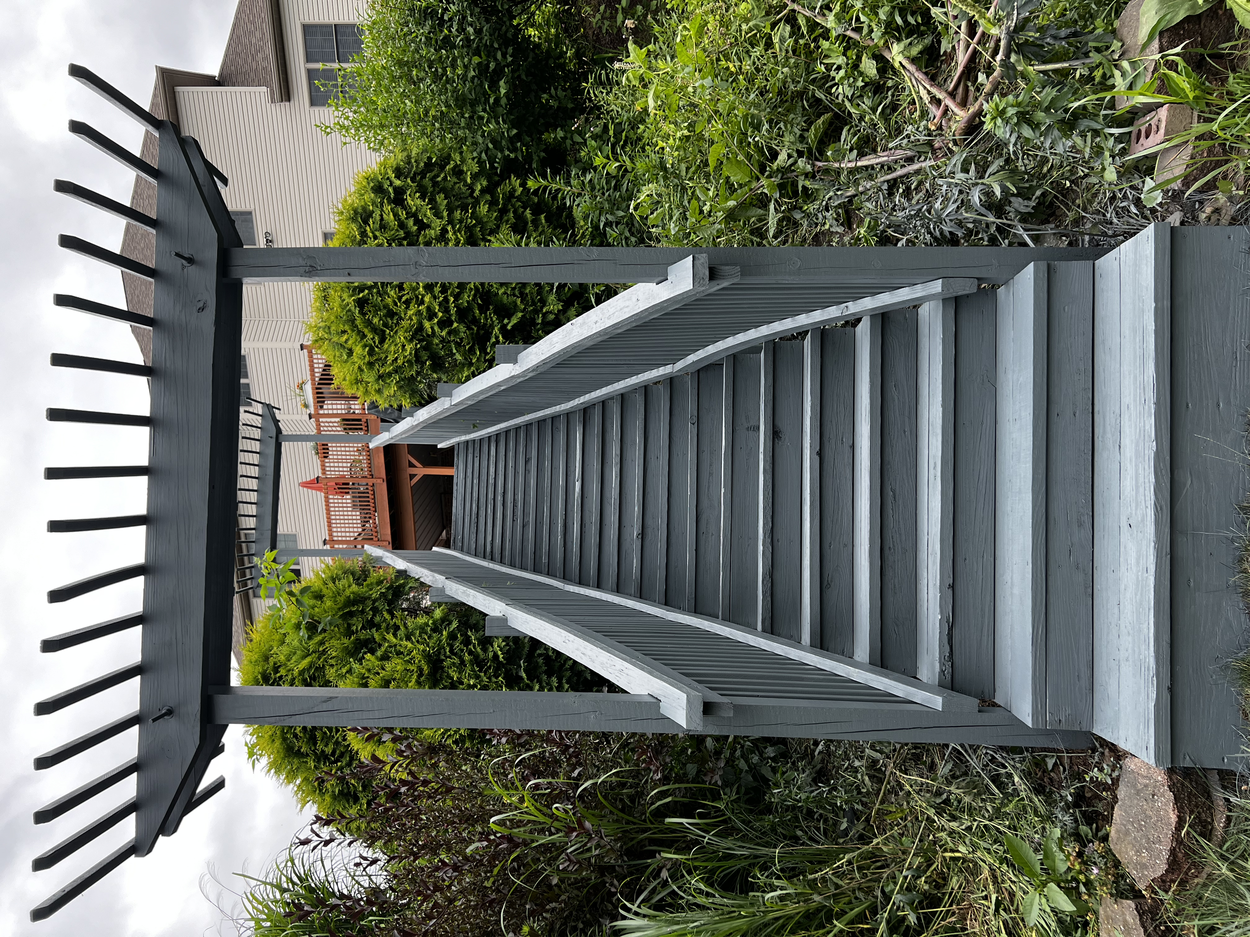 Backyard deck with gray painted wood steps and a black garden trellis or arbor at the top, surrounded by green bushes and plants.