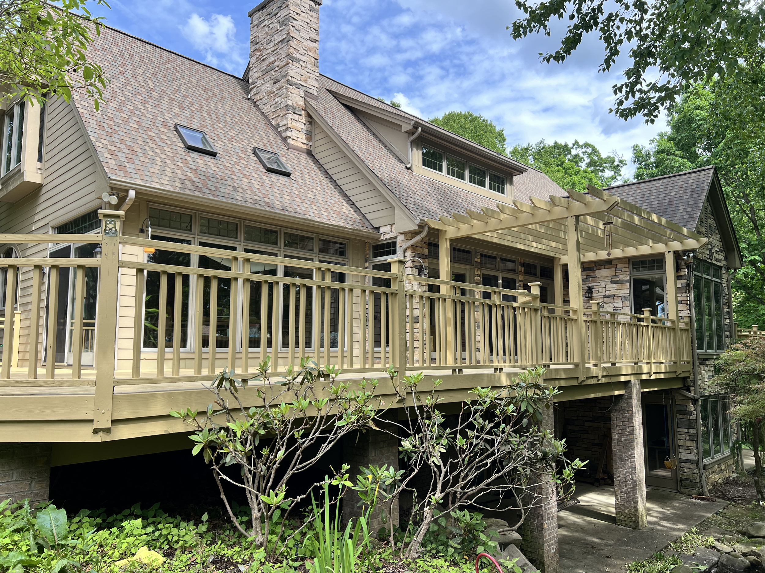 Back of a house with a deck, beige railing, windows, and a stone chimney, surrounded by trees and greenery under a partly cloudy sky.
