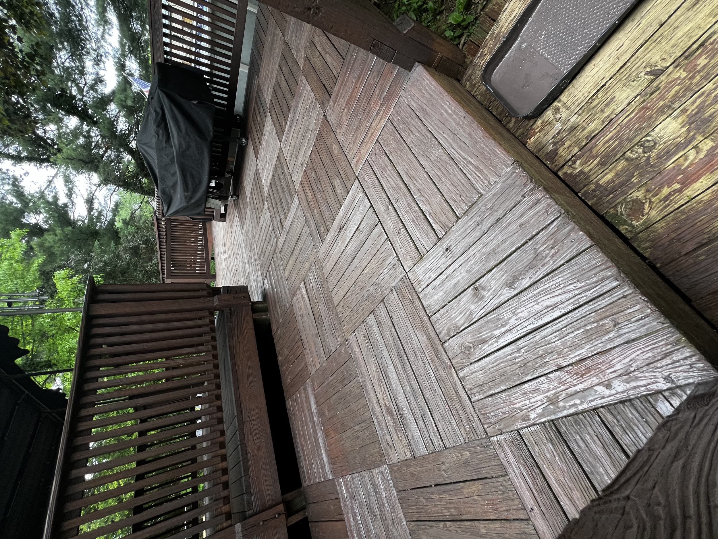A wooden deck with a built-in grill covered by a black grill cover, surrounded by a wooden railing, with trees and greenery in the background.