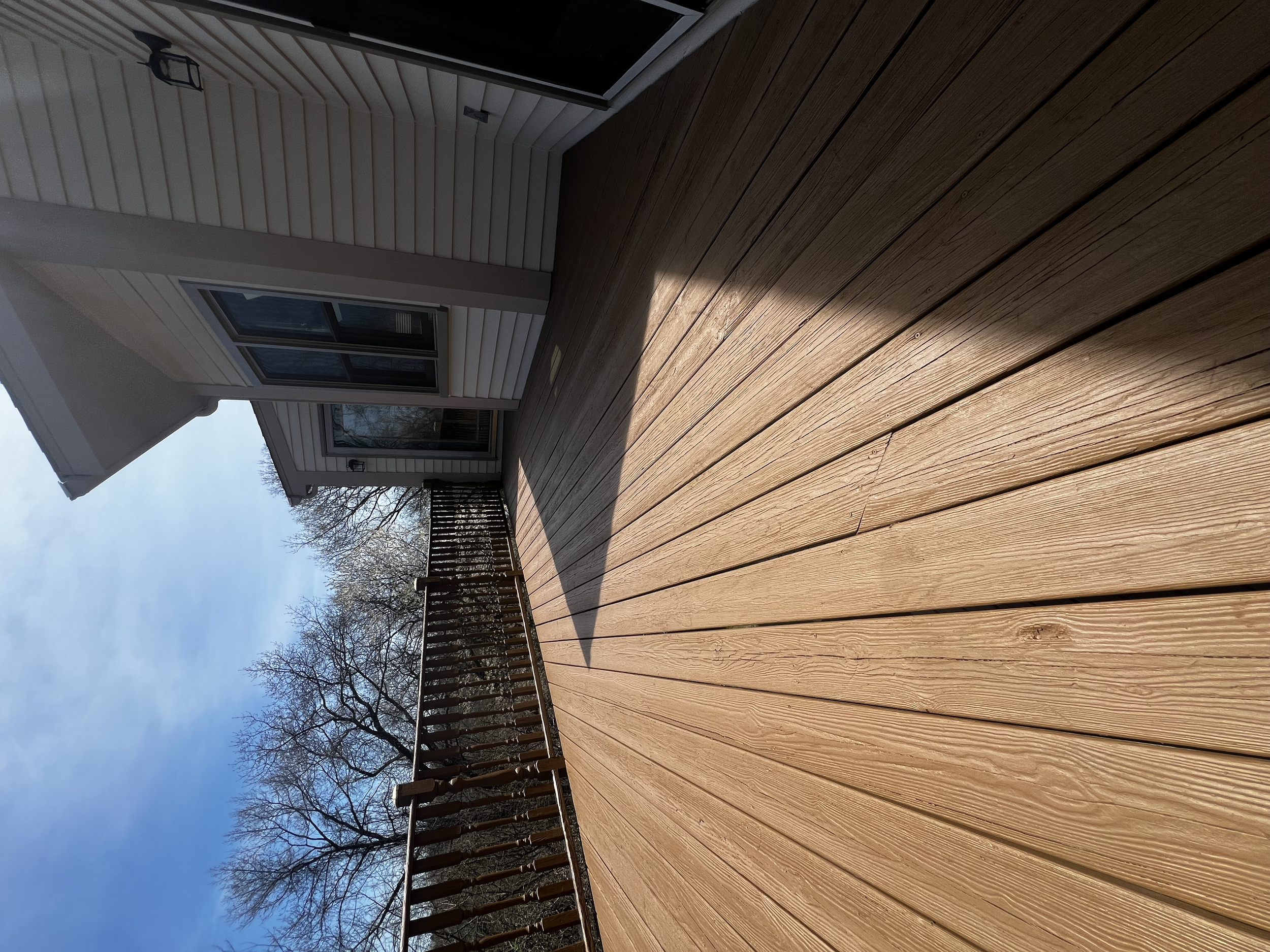 A wooden deck with a railing, attached to a house with white siding and a window, under blue sky with leafless trees in the background.