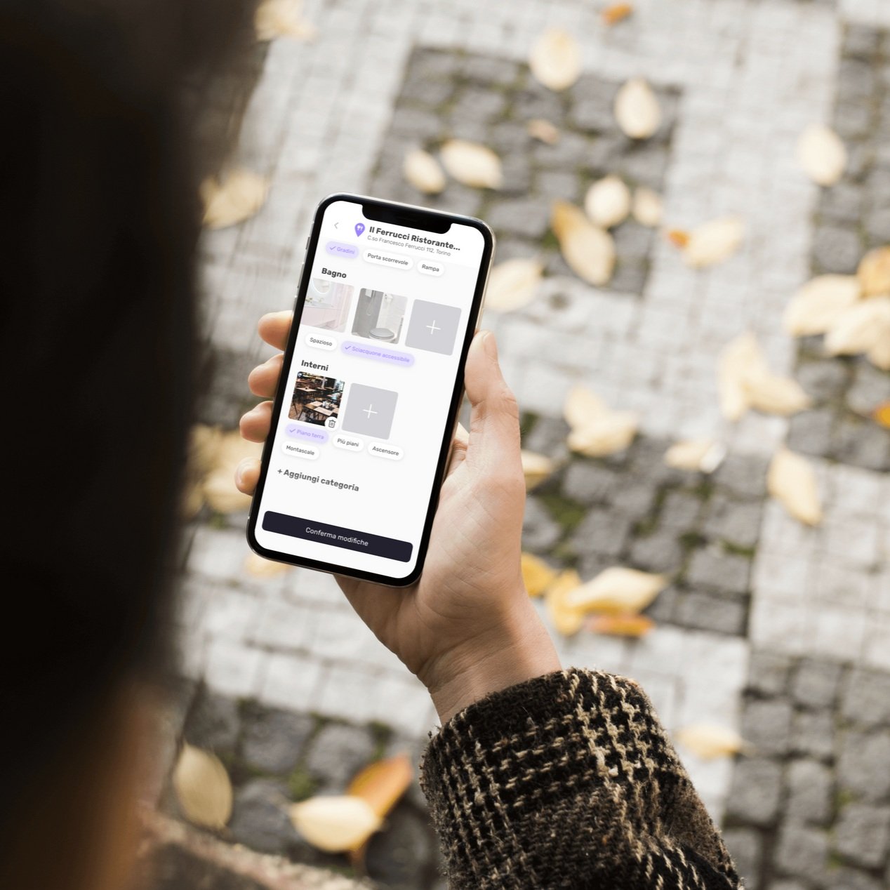 Person holding a smartphone displaying an app interface with options for adding or selecting categories, such as bathroom and interior. Background shows a tiled pavement with fallen leaves.