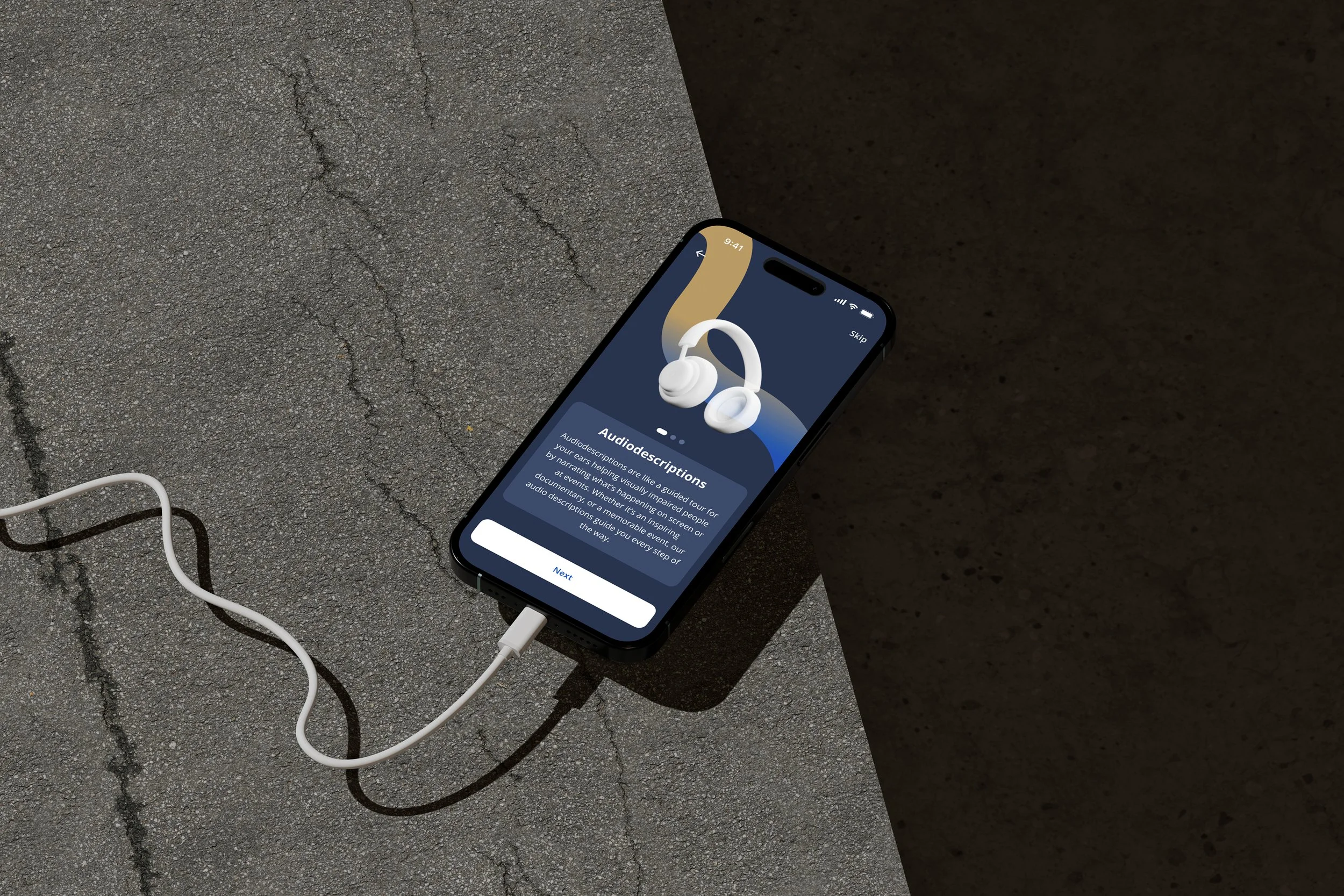 Smartphone on asphalt showing audio description setup screen with headphones image and charging cable attached.