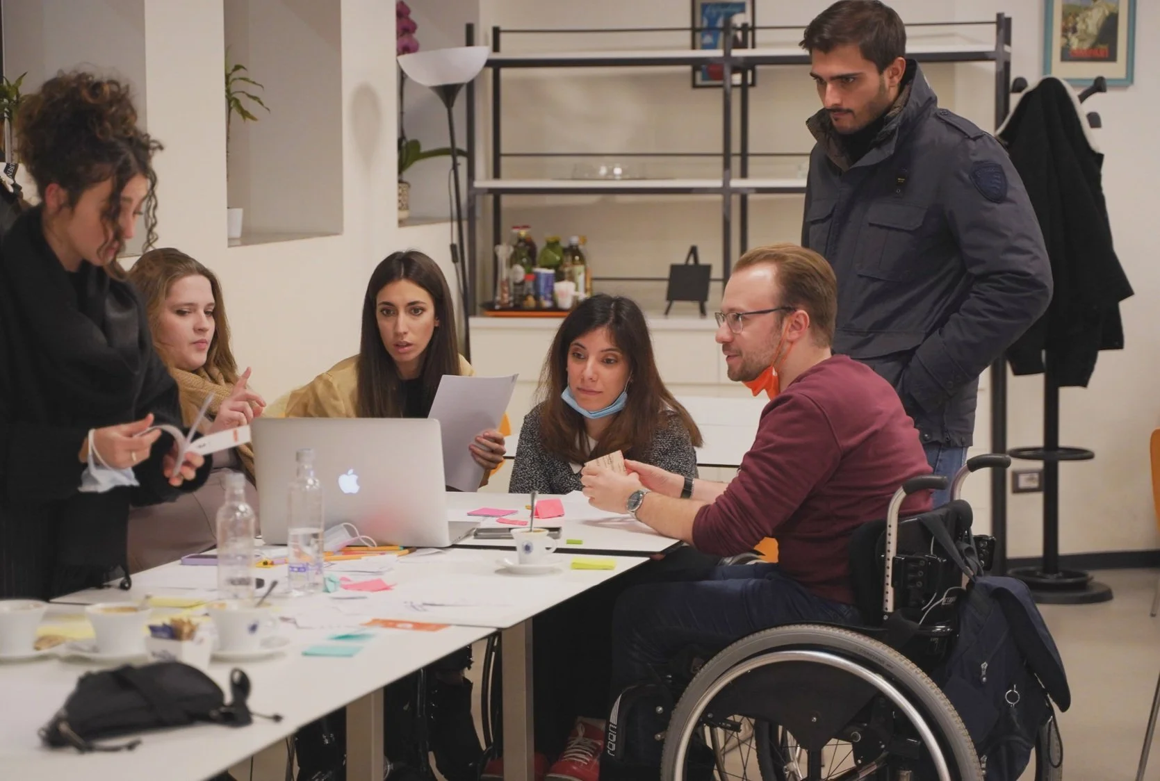 Group of people having a discussion around a table with a laptop, papers, and sticky notes in an office setting.