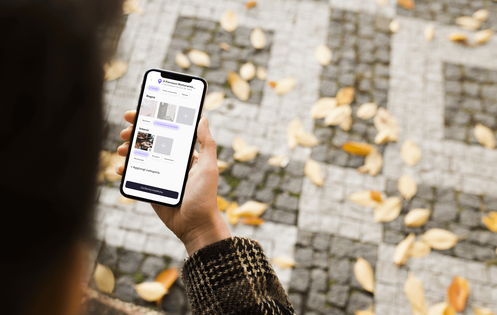 Person holding a smartphone displaying an app interface with categories like bathroom and interiors, standing on a cobblestone path with scattered autumn leaves.