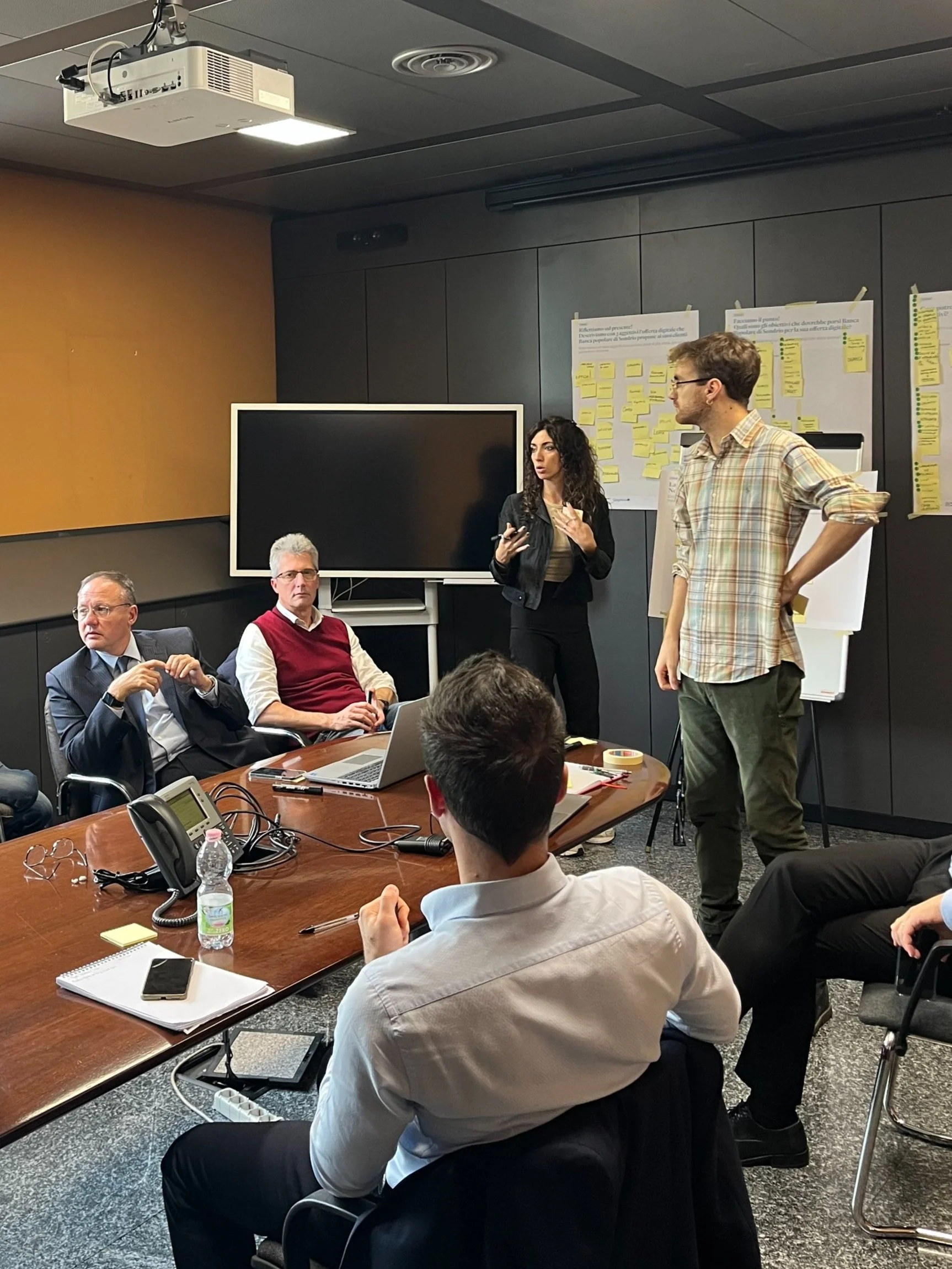 Workshop with several people around a conference table. A man and a woman are standing, presenting. Others seated are listening, with a large display and papers in the background.