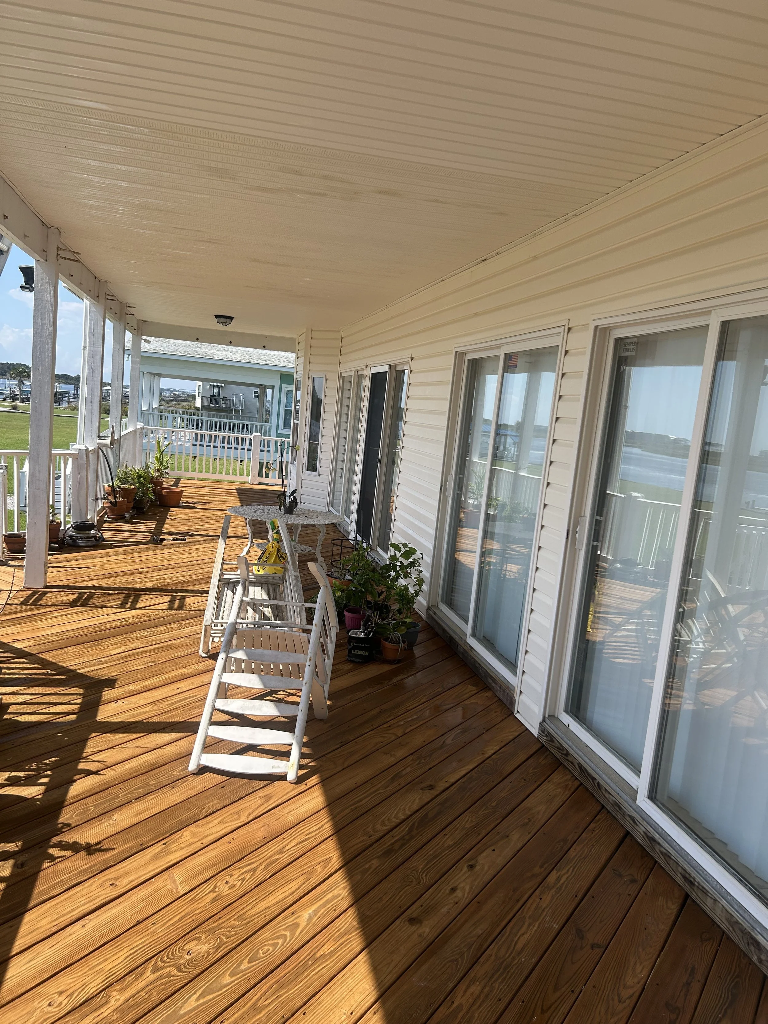 A spacious porch with wooden flooring, white railing, and sliding glass doors leading indoors. Several potted plants and outdoor furniture are visible, with clear skies and water in the background.