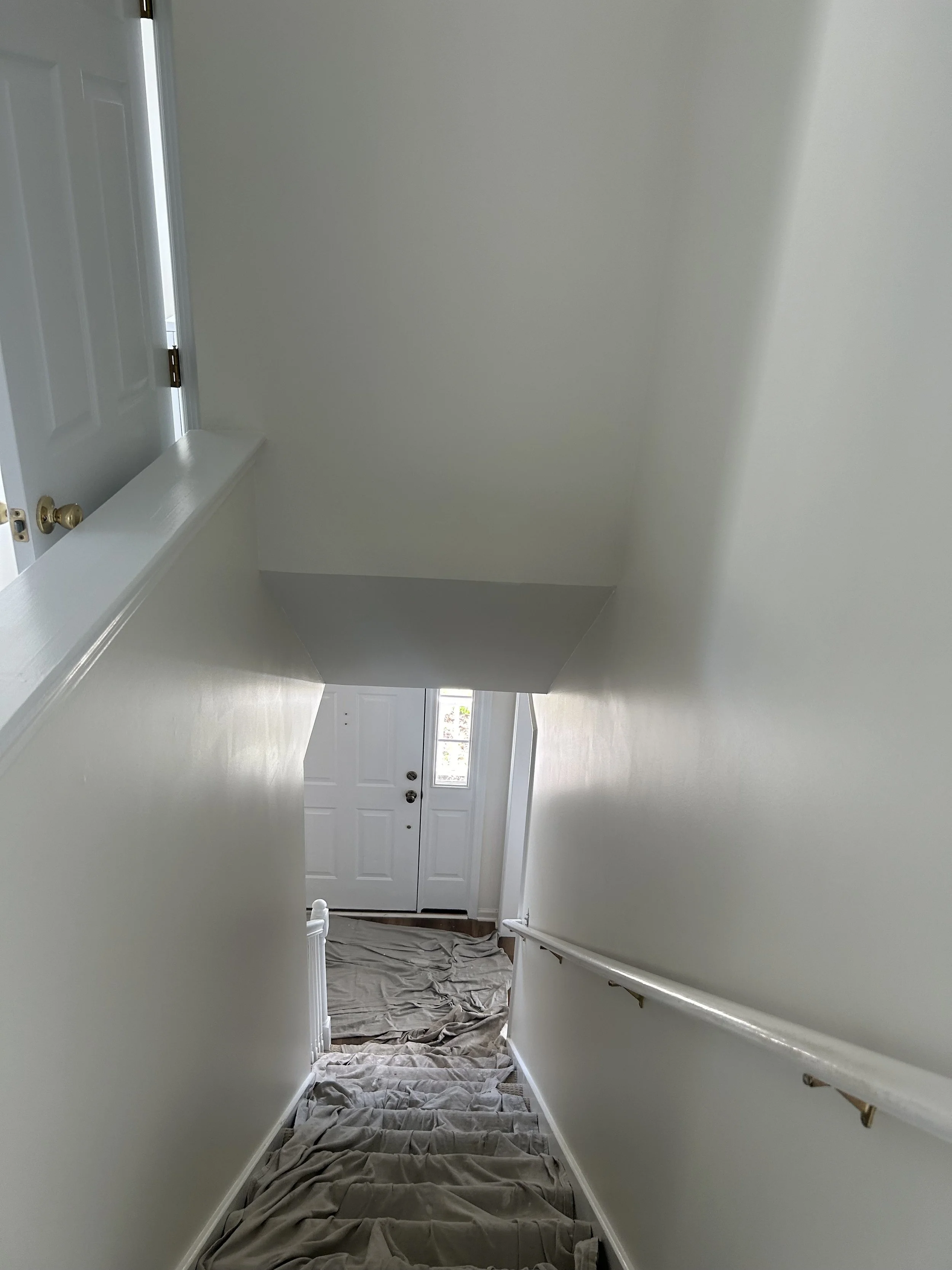 Empty staircase with beige carpet, white walls, and a white door at the bottom, partially covered by drop cloths.