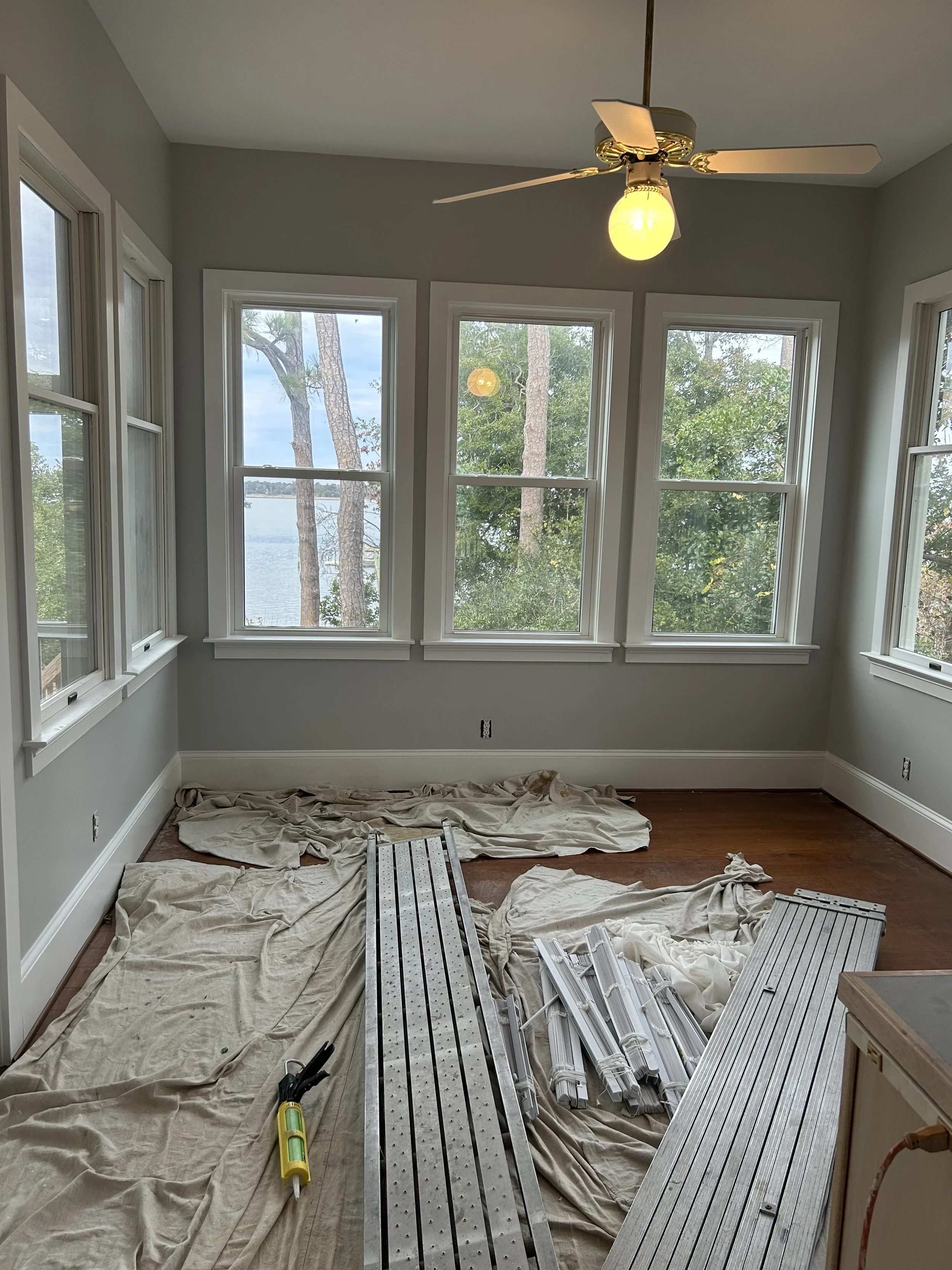 Room with multiple large windows overlooking trees and water, ceiling fan with light, flooring in progress, tools and construction materials on the floor covered with cloths.