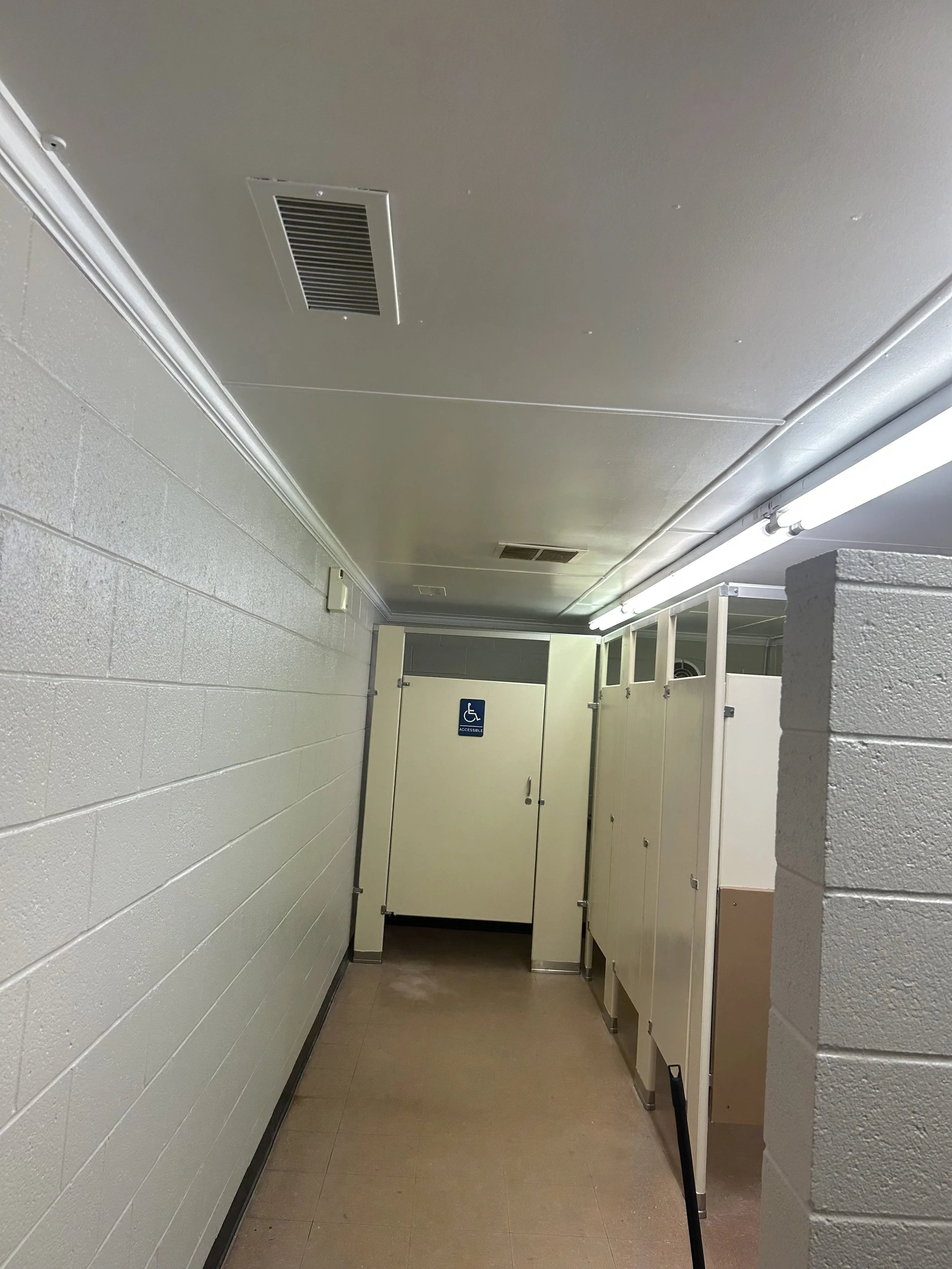 Public restroom with accessible stall and urinals, ceiling vents, fluorescent lighting, and beige tile floor.